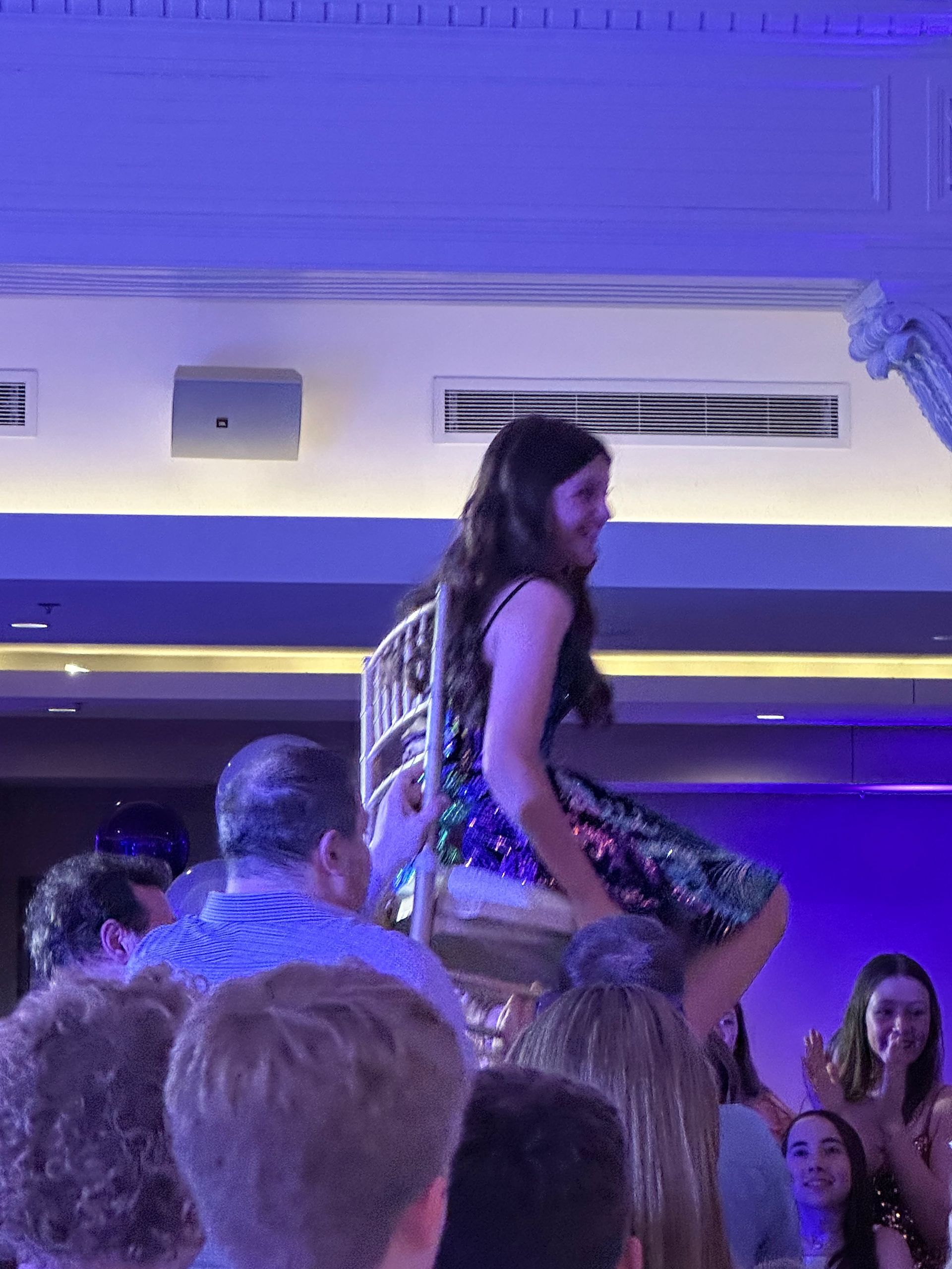Teen girl smiles while lifted in a chair at a party, surrounded by people, under blue and white lights.