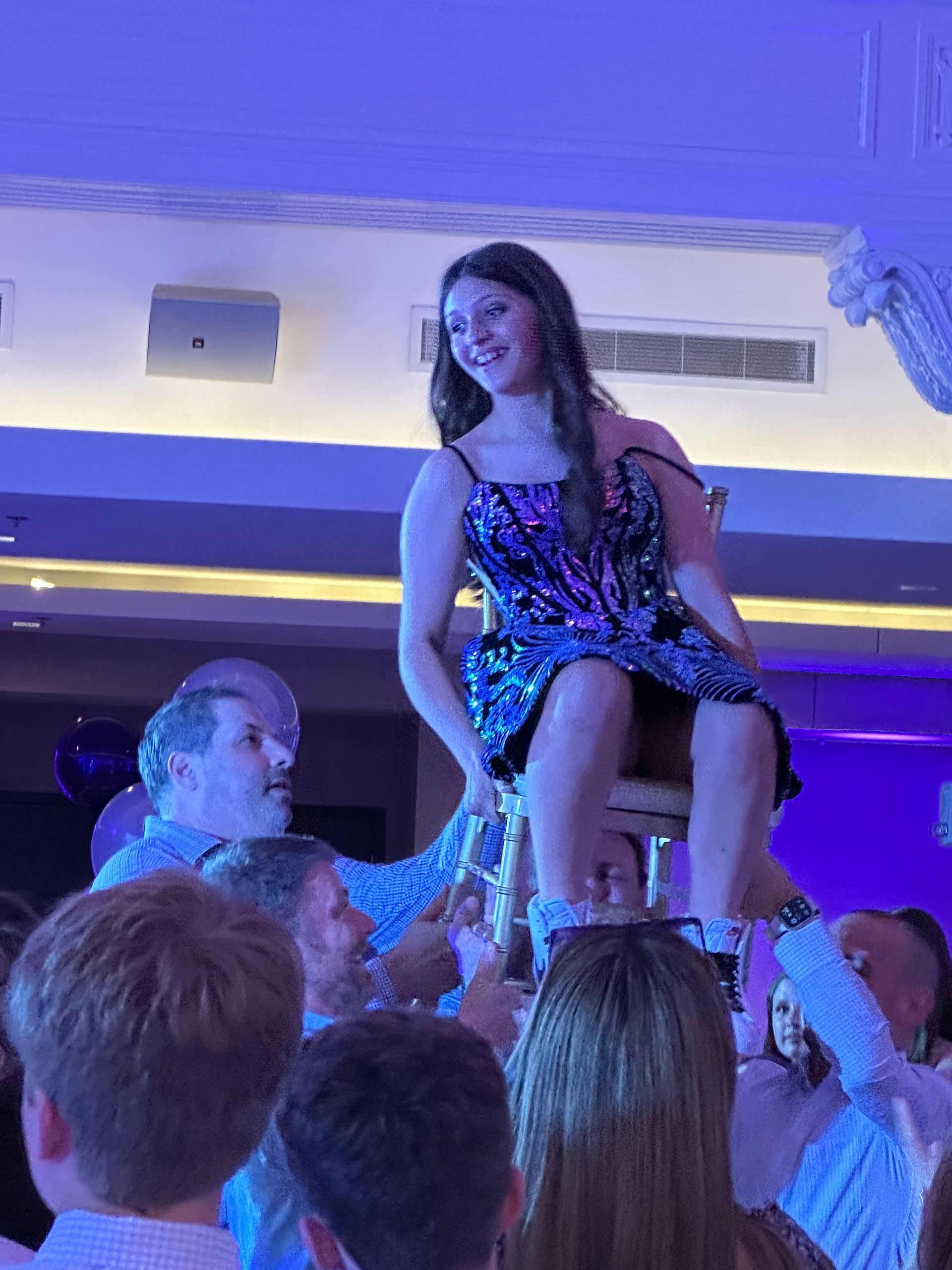 A woman in a sparkly dress is lifted on a chair by a crowd at a party.