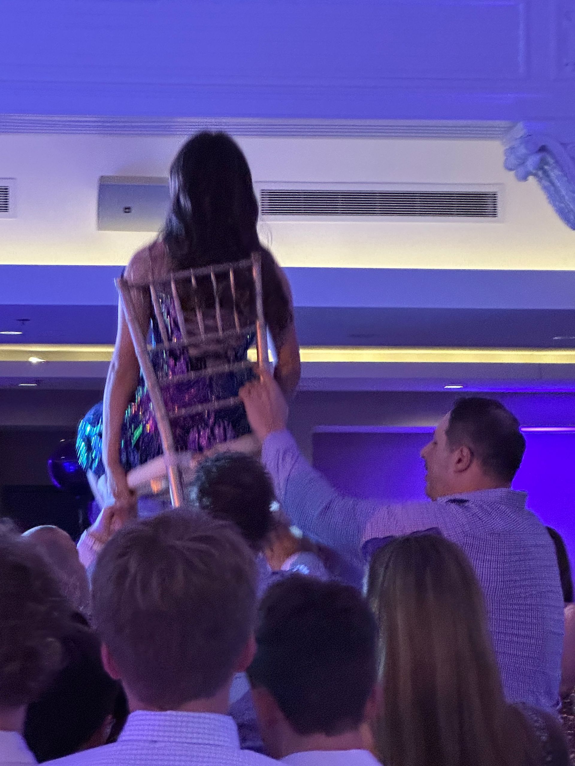 People lifting a person seated on a chair at a celebration; woman in sparkly dress.