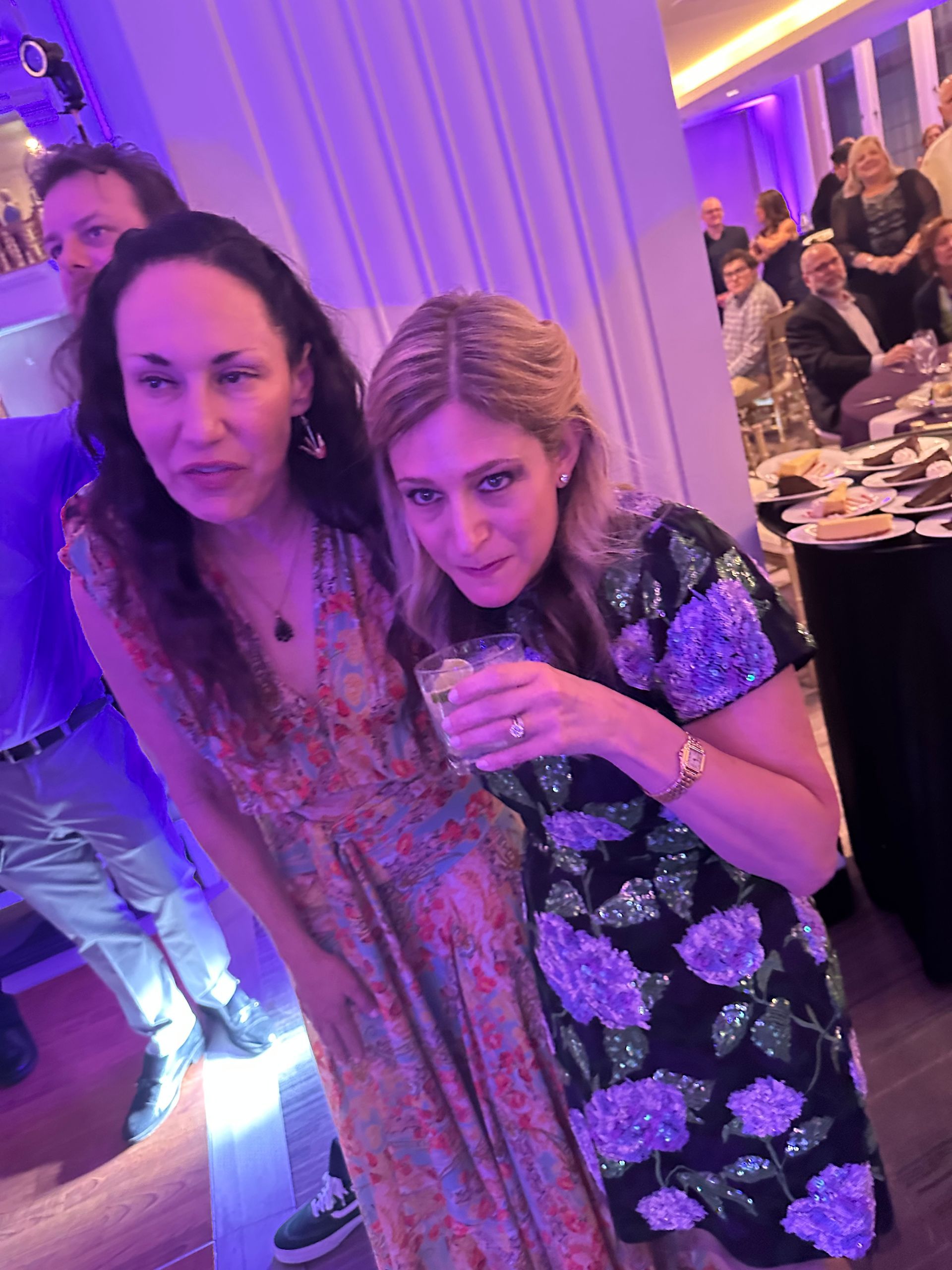 Two women at a party; one drinks, the other looks on. Purple-lit room, others in background.
