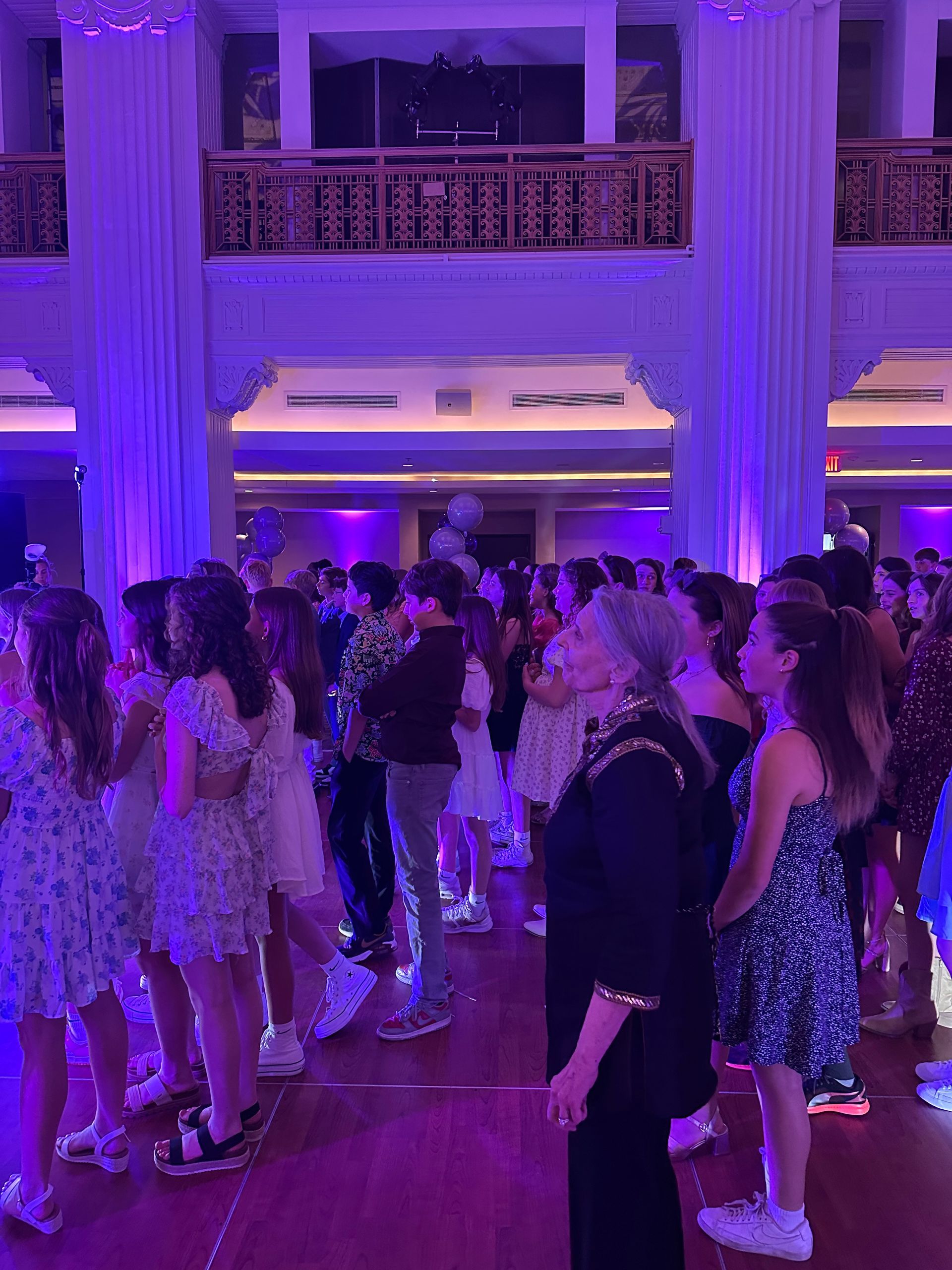 People at a party in a large ballroom with purple lighting; many are young women in dresses.