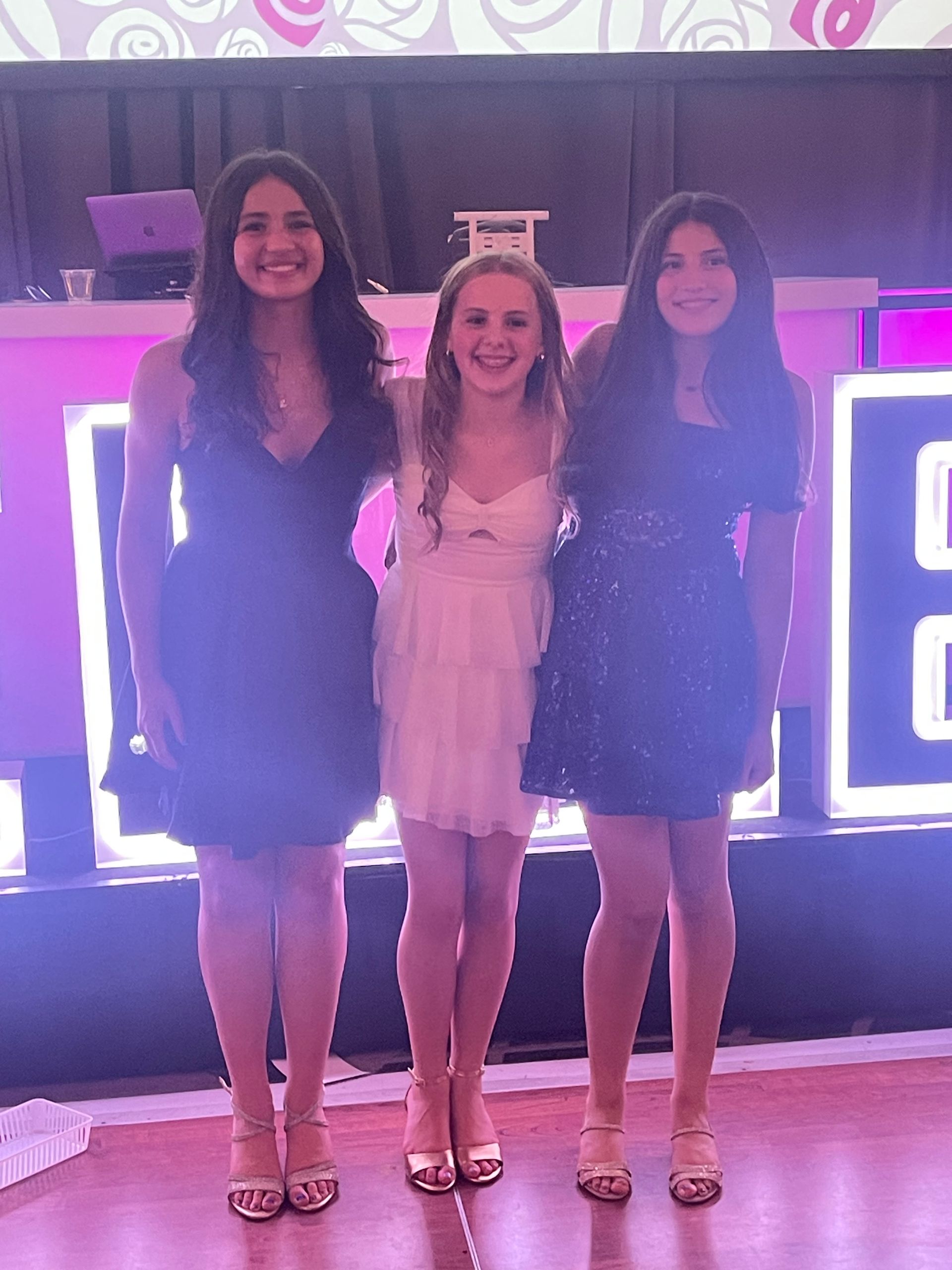 Three young women pose together at an event, wearing dresses and heels.