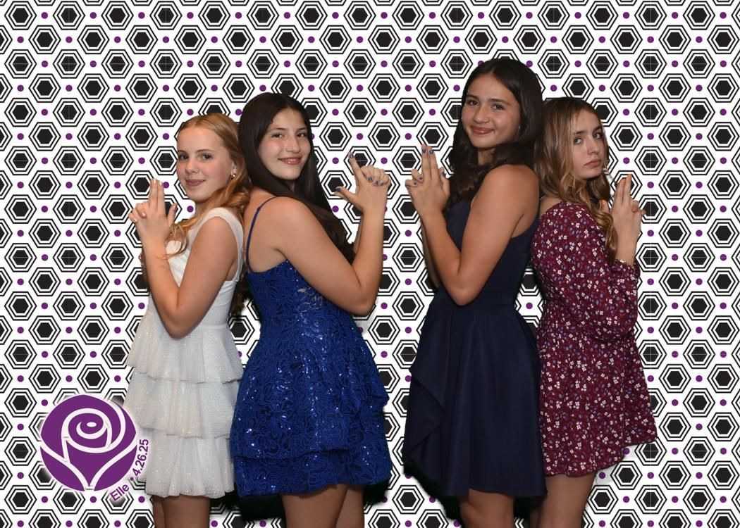 Four young women in formal dresses pose back-to-back, making gun-like hand gestures in front of a patterned backdrop.