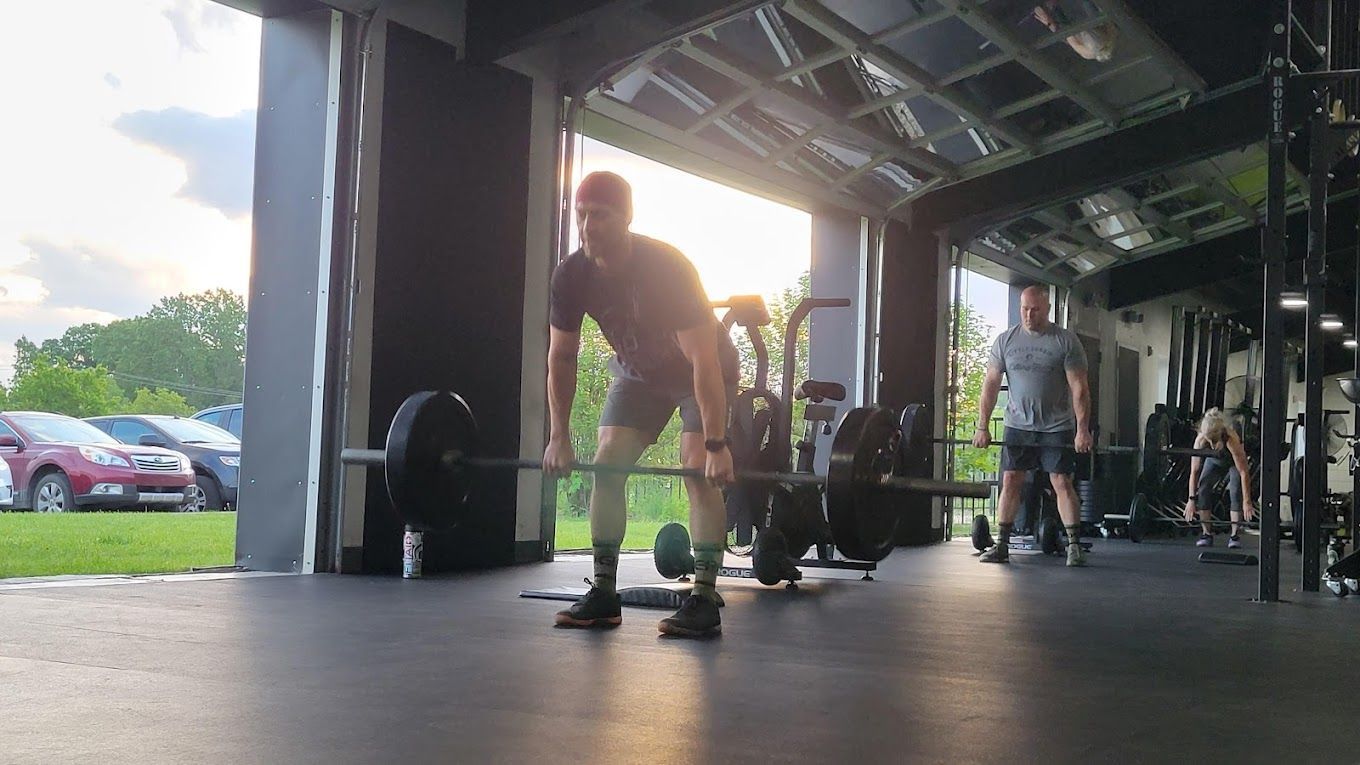 Man lifting a barbell in a gym with open doors, other people in the background.
