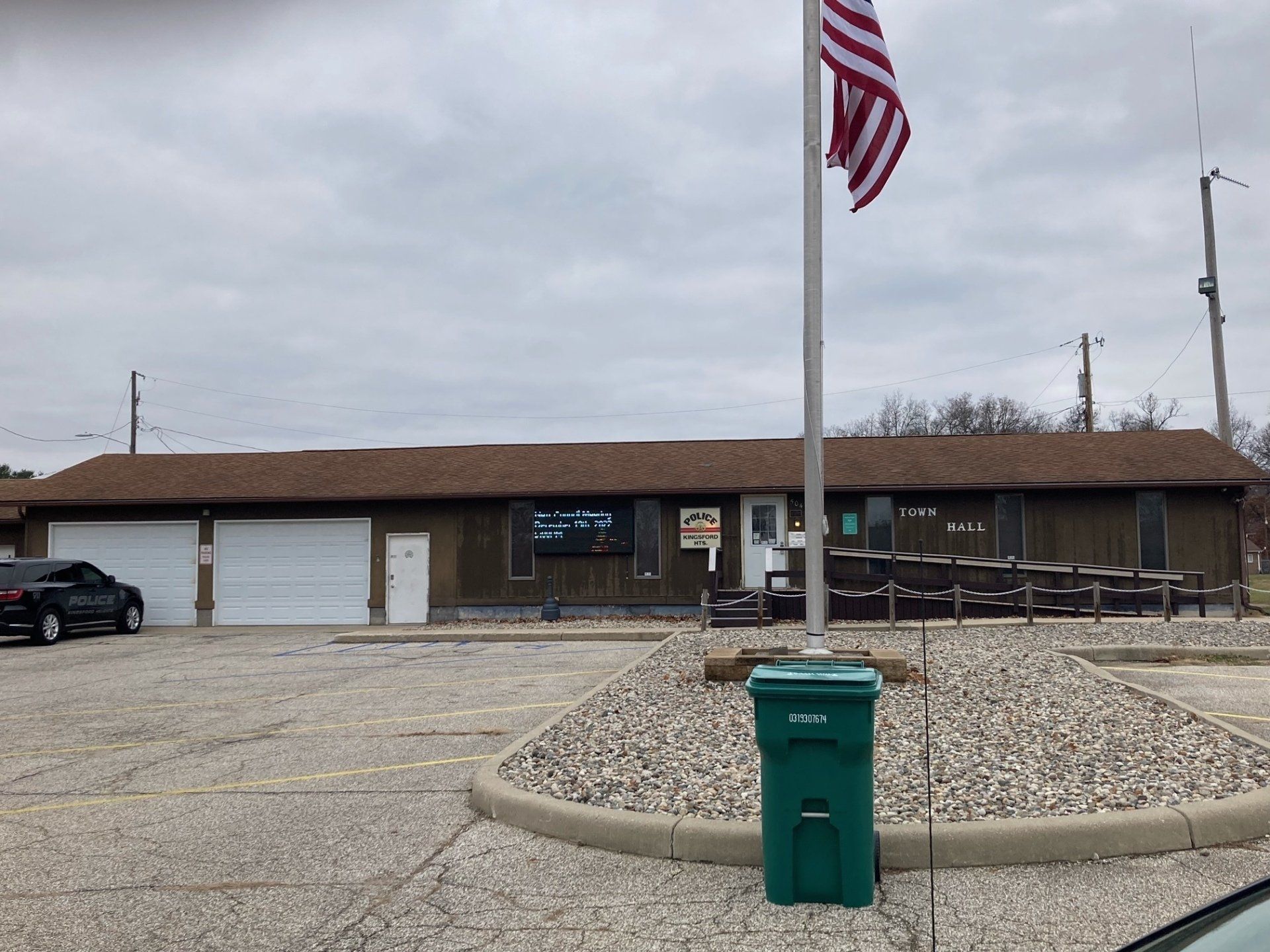 A green trash can is in front of a building with a flag on a pole.