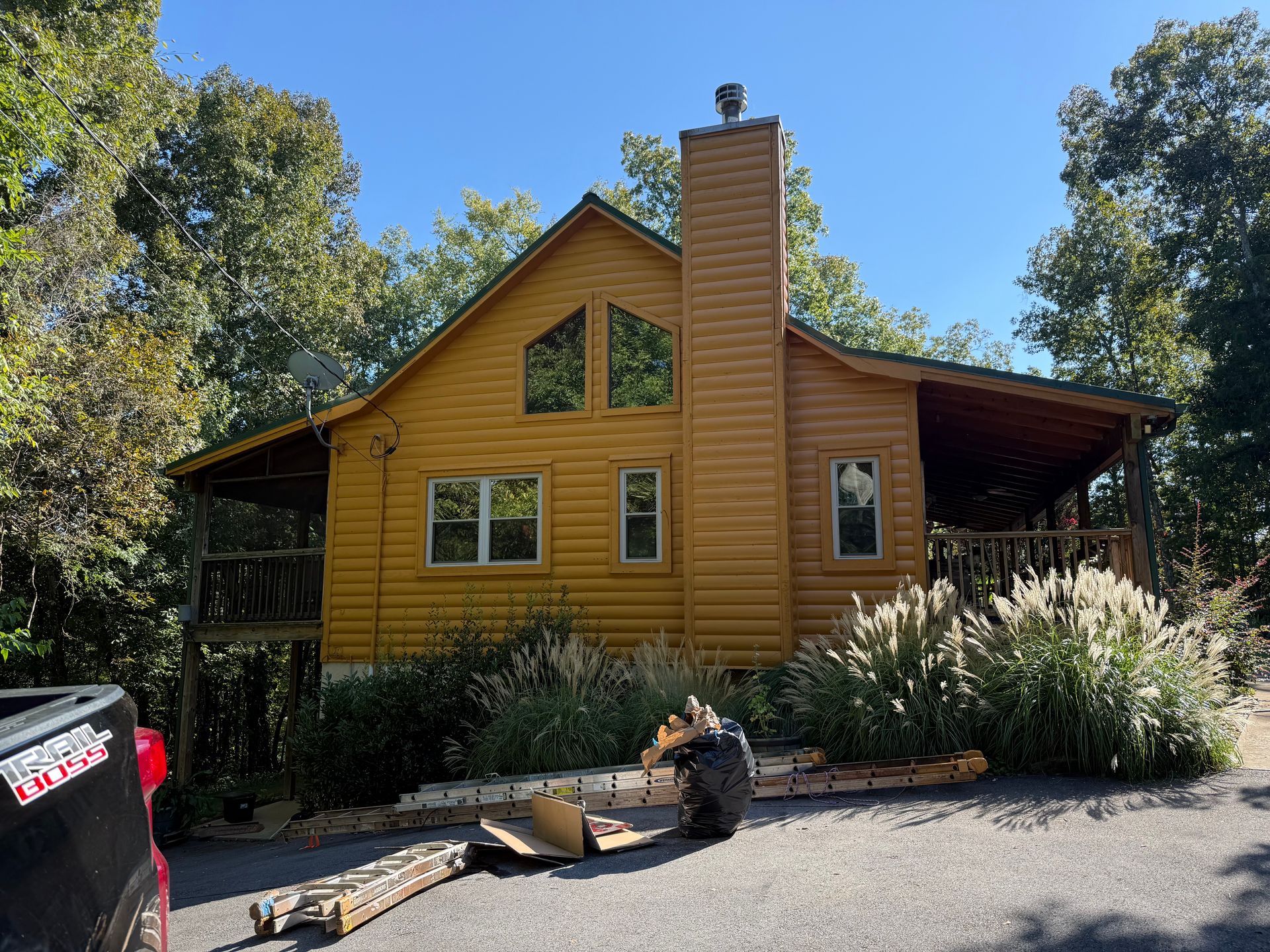 Yellow cabin with chimney, porch, and landscaping under a blue sky, surrounded by trees.