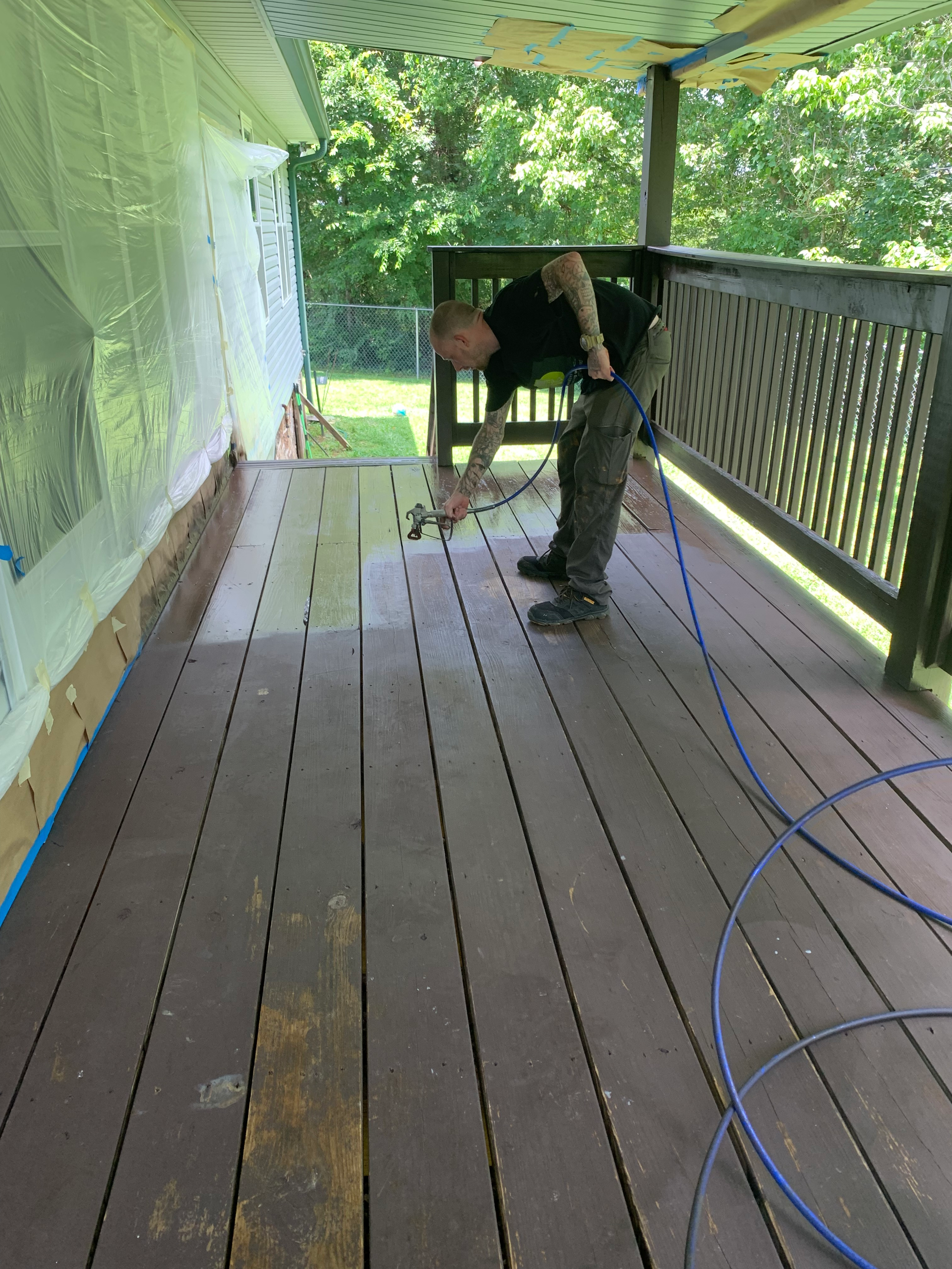 Person spray painting a wooden deck with a sprayer; house in the background.