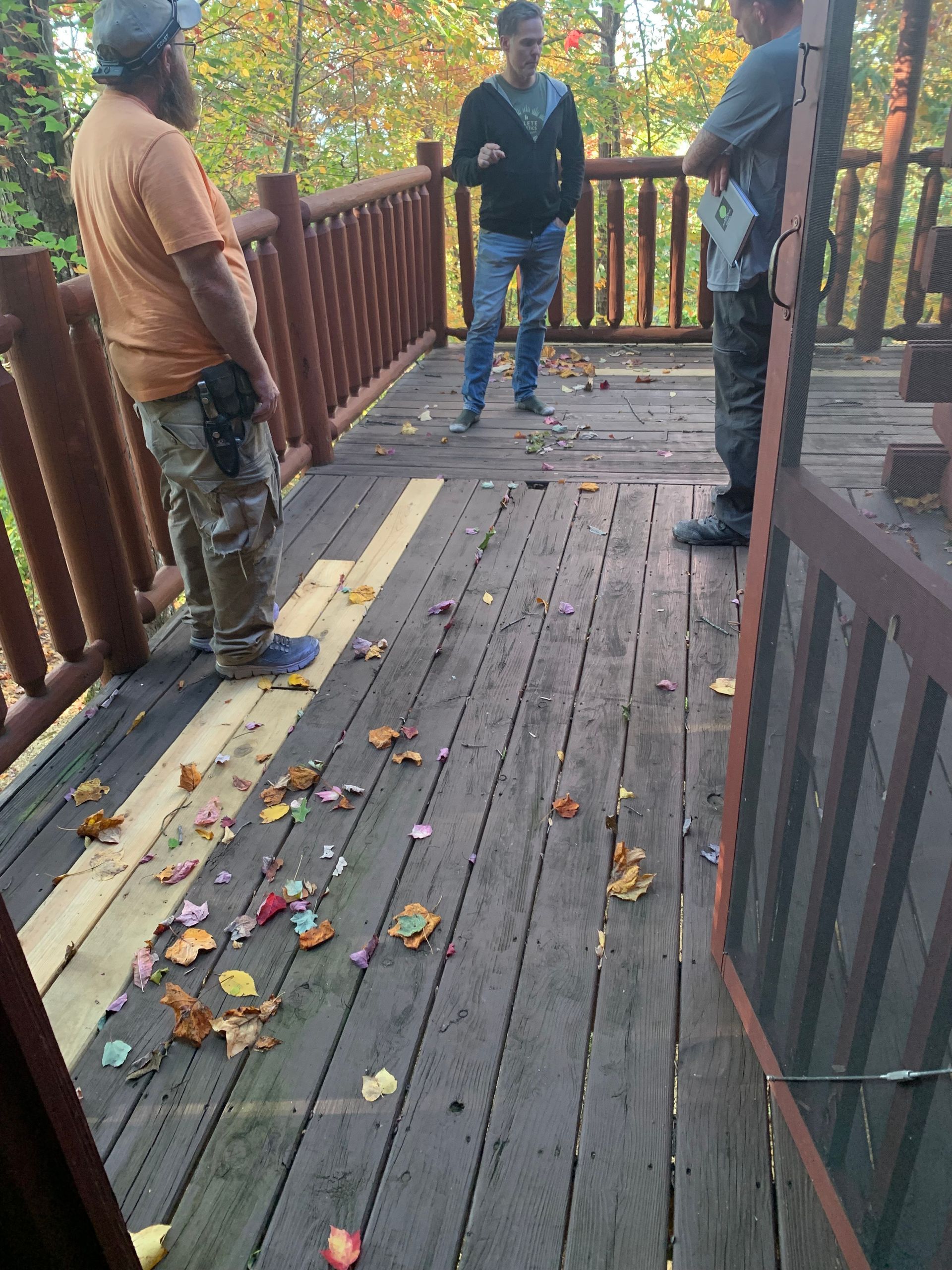 Three people on a wooden deck with railing, discussing deck repair. Leaves are scattered.