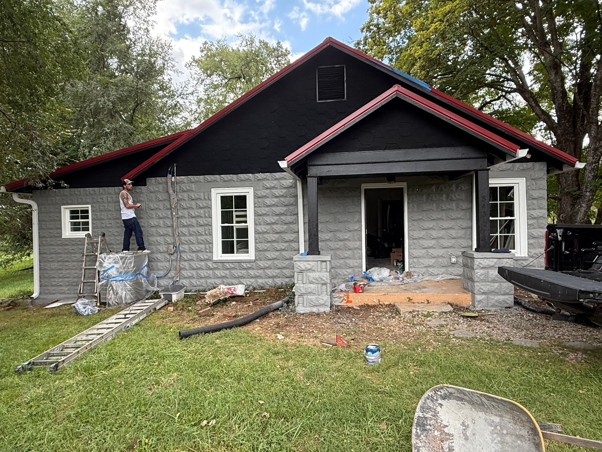 Home exterior under construction: gray brick, black roof, man on ladder, materials on ground.