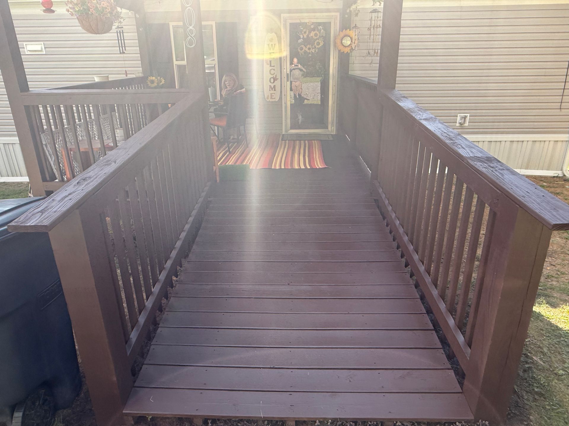 Wooden ramp leading to a doorway, brown railings, red rug in front of door, sunlight.