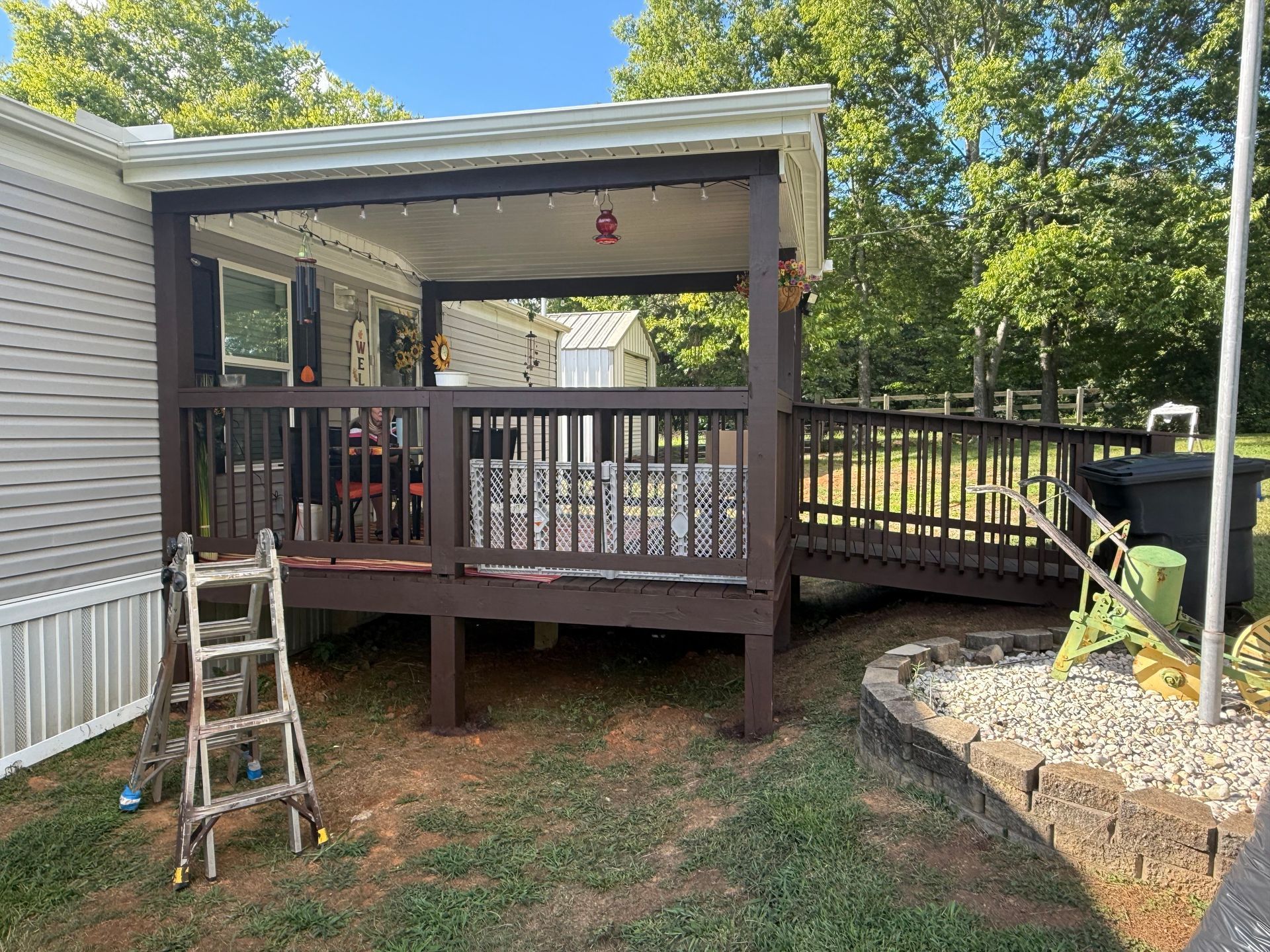 Brown deck with ramp attached to a mobile home, ladder in foreground, sunny day.