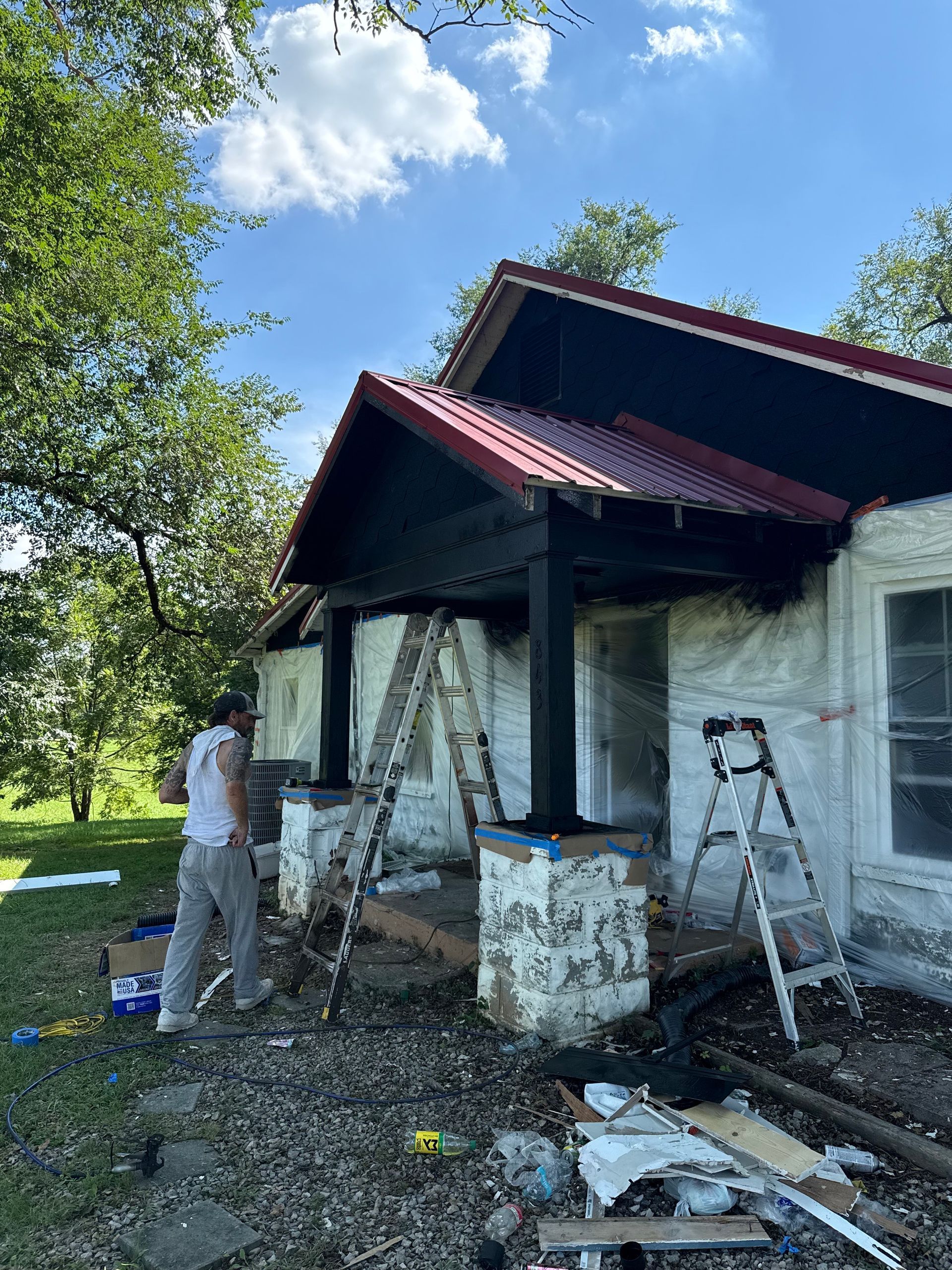 Person painting porch columns black on a house with a red metal roof. Ladders and construction supplies are present.