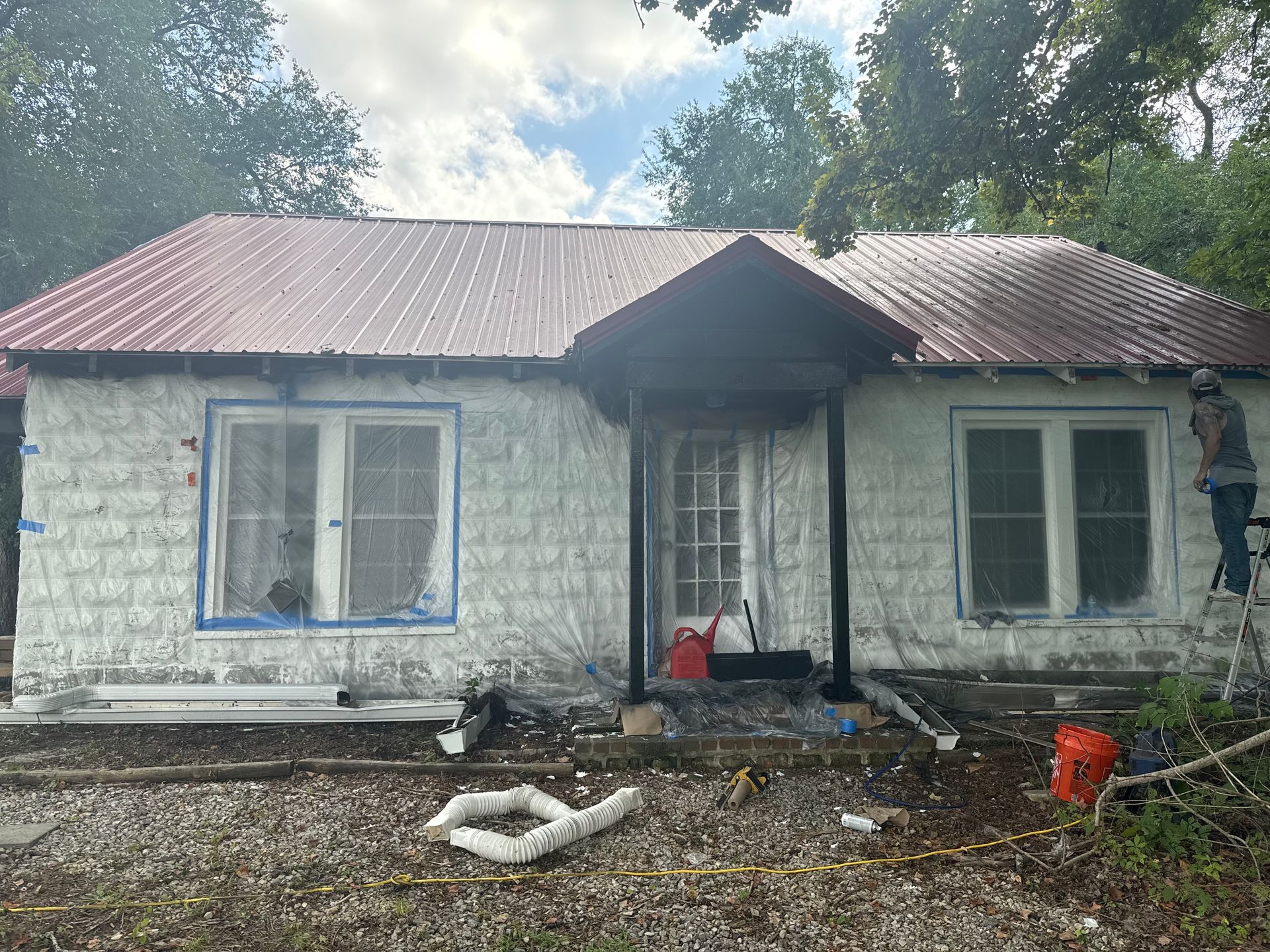 House exterior being painted, covered in white wrap, with red metal roof. A person on a ladder.