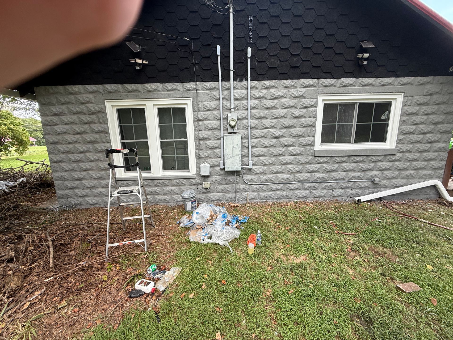 Gray brick building exterior with electrical conduit and windows; ladder and debris on grass.