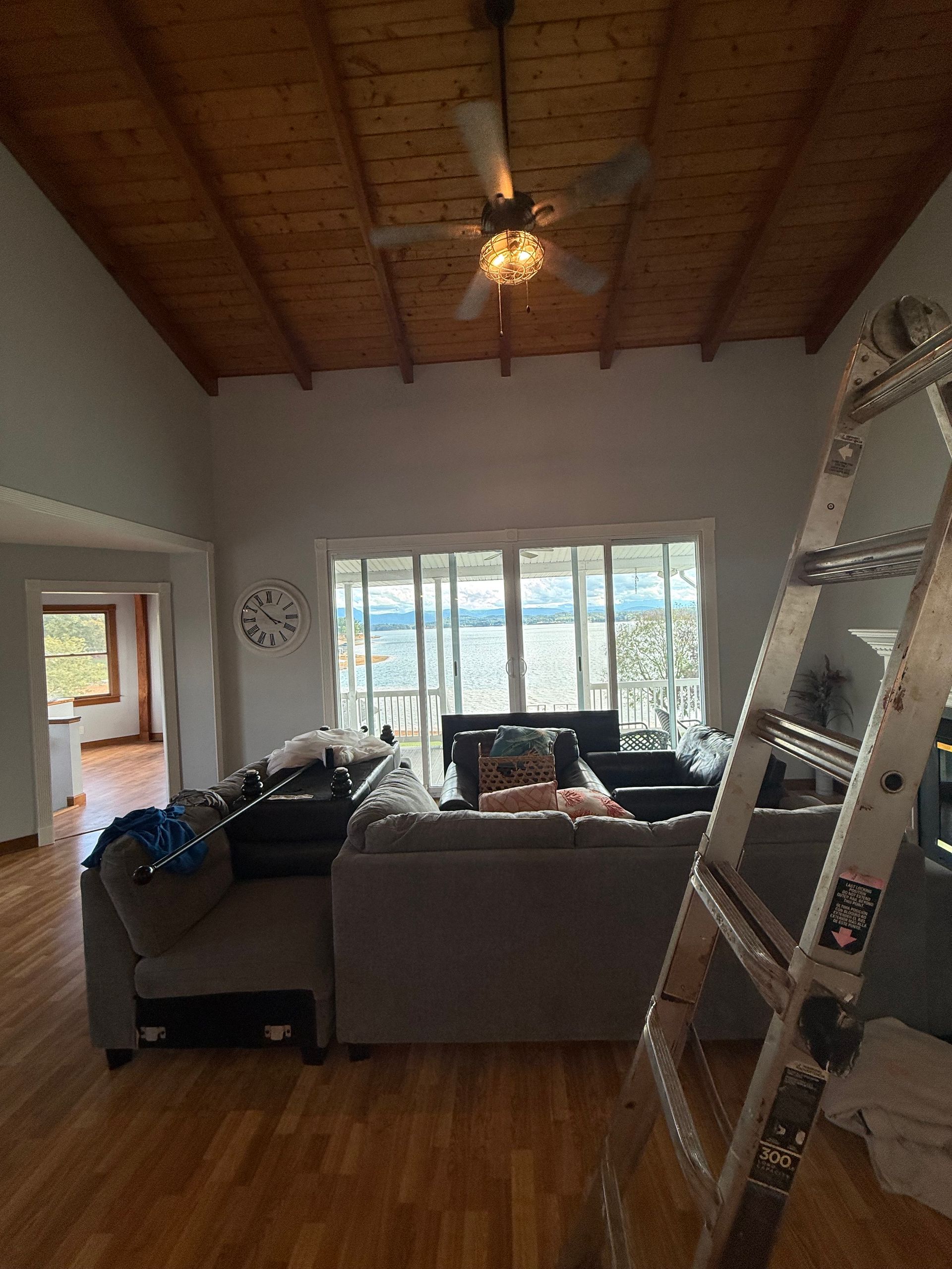 Living room with a high wood ceiling, gray sectional sofa, and a ladder. Large windows show an outdoor view.