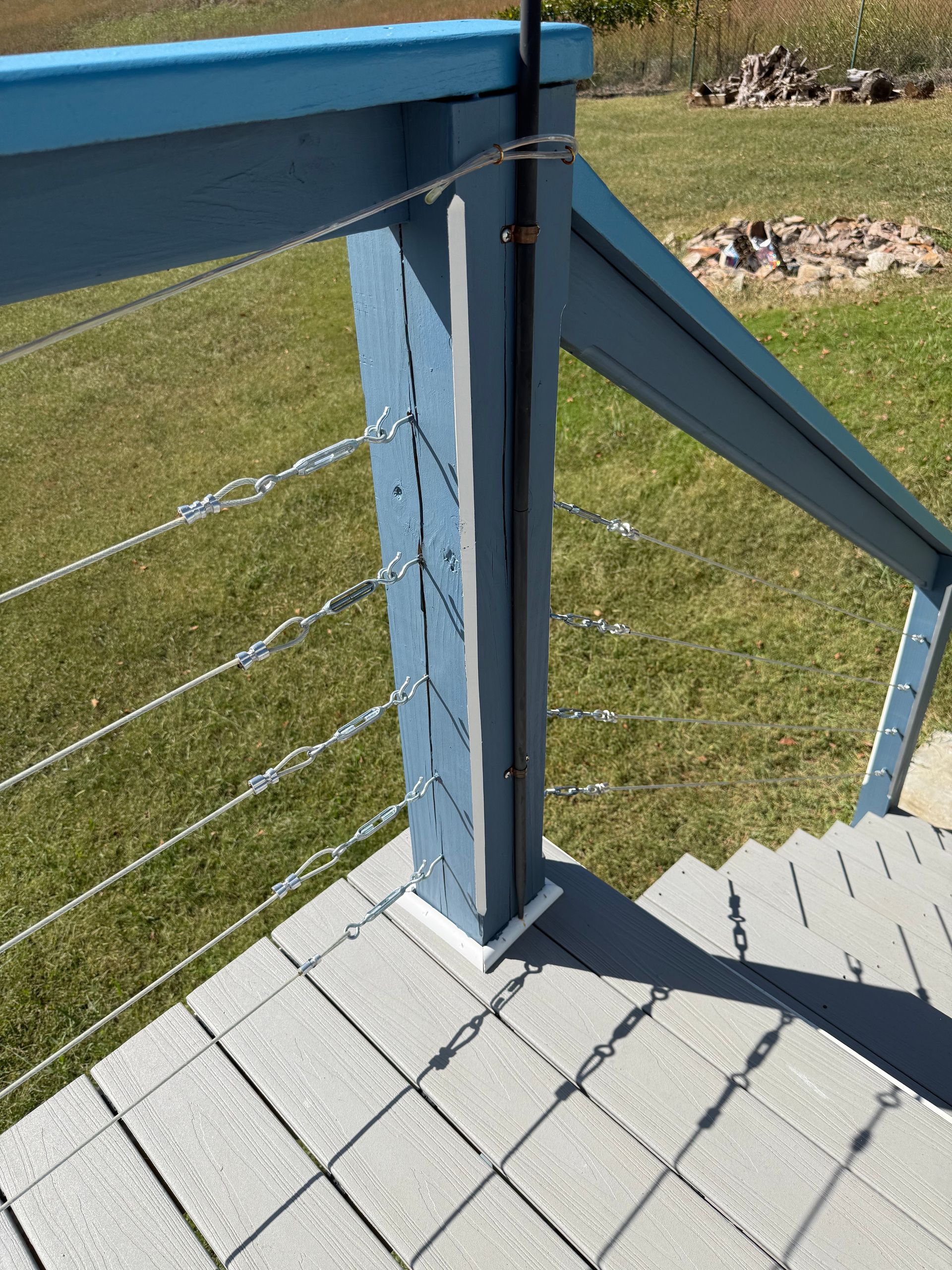 Blue deck railing with cable wires, on gray tiled deck. Green grass in background.