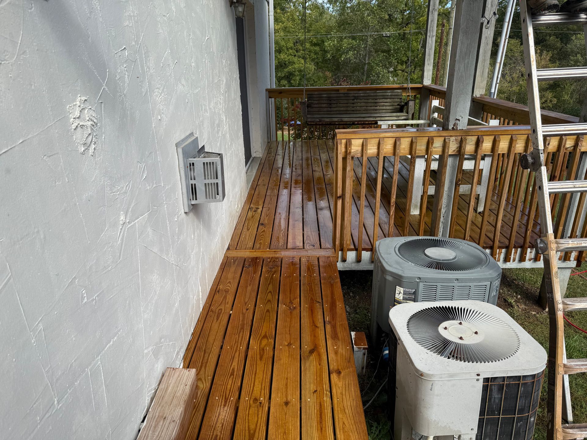 Wooden deck next to a white stucco wall, two AC units. A ladder is propped up on the deck.