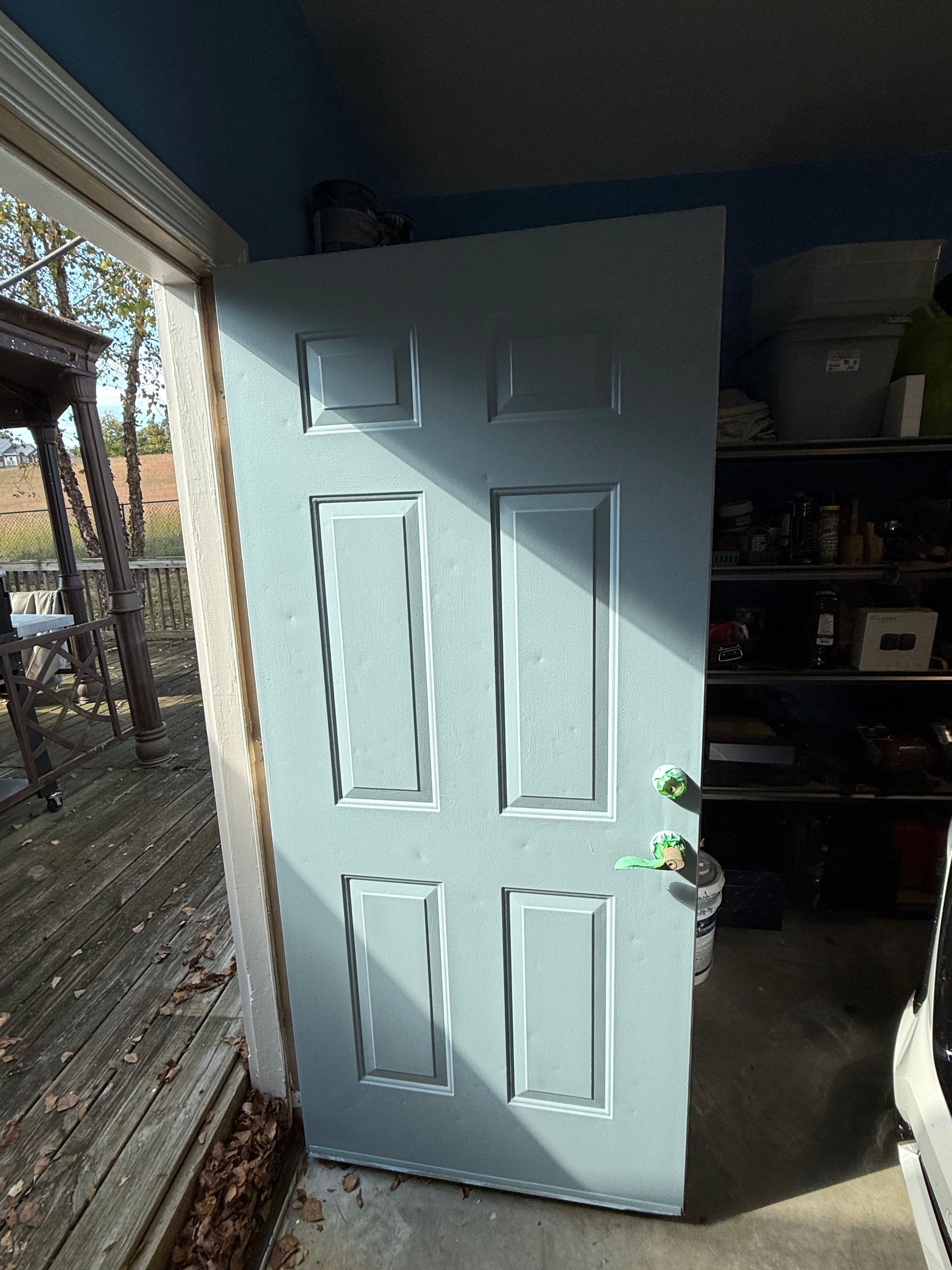 Blue exterior door slightly ajar, casting shadows. Wooden deck to the left, shelves in garage to the right.