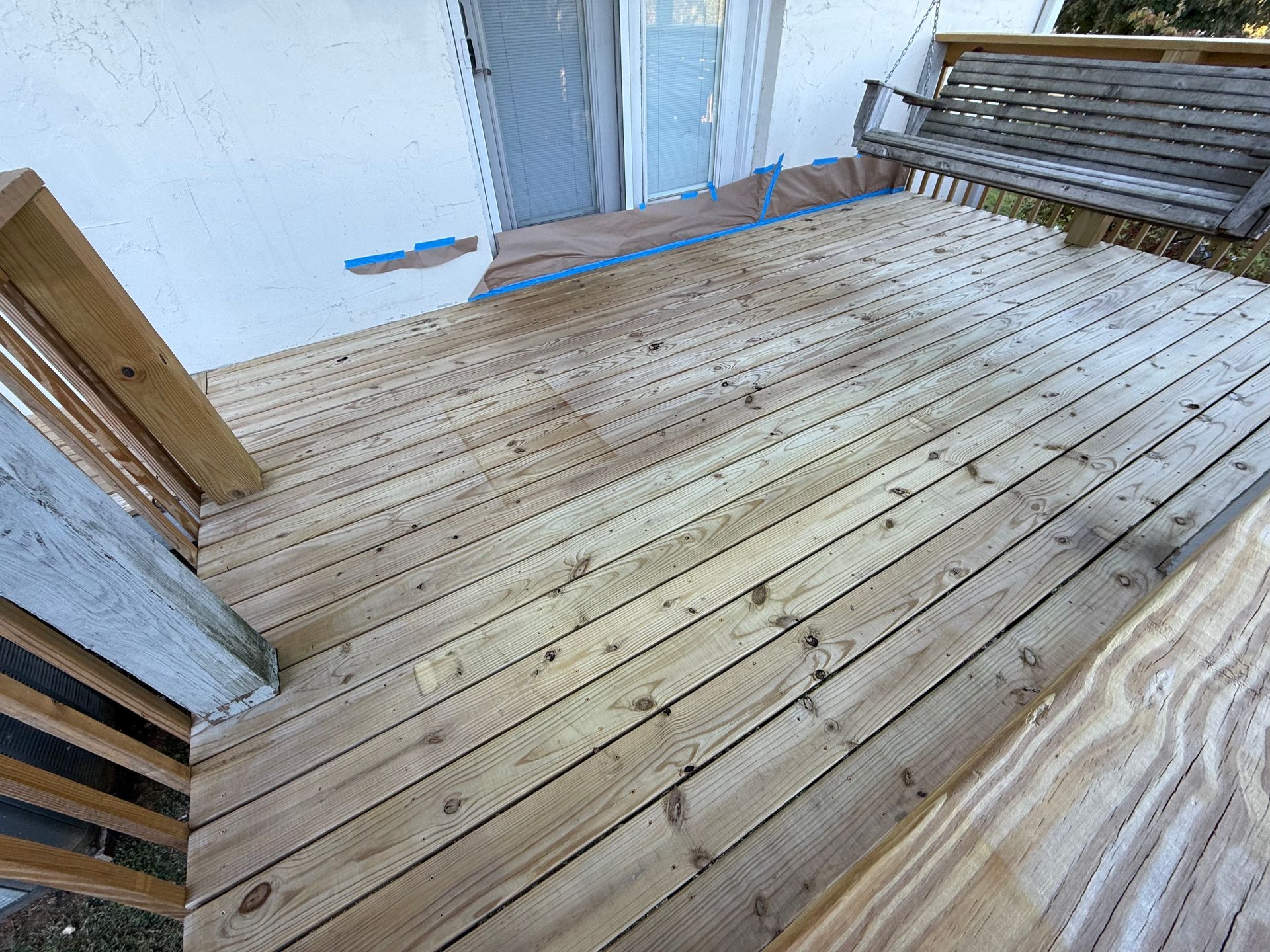 Wooden deck with a porch swing, next to a white house with a door.