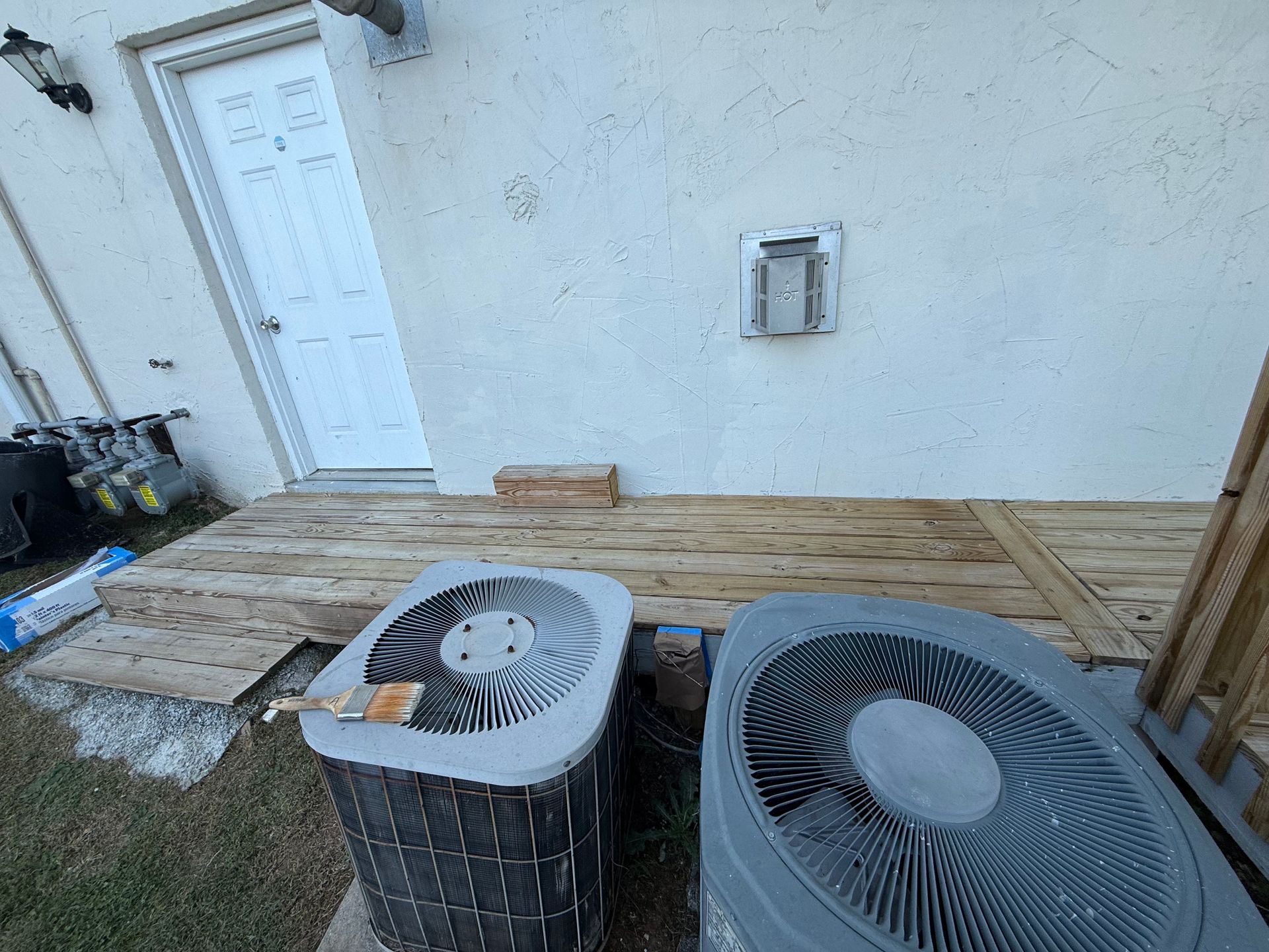 Two air conditioning units on a wooden deck next to a white stucco wall and a white door.