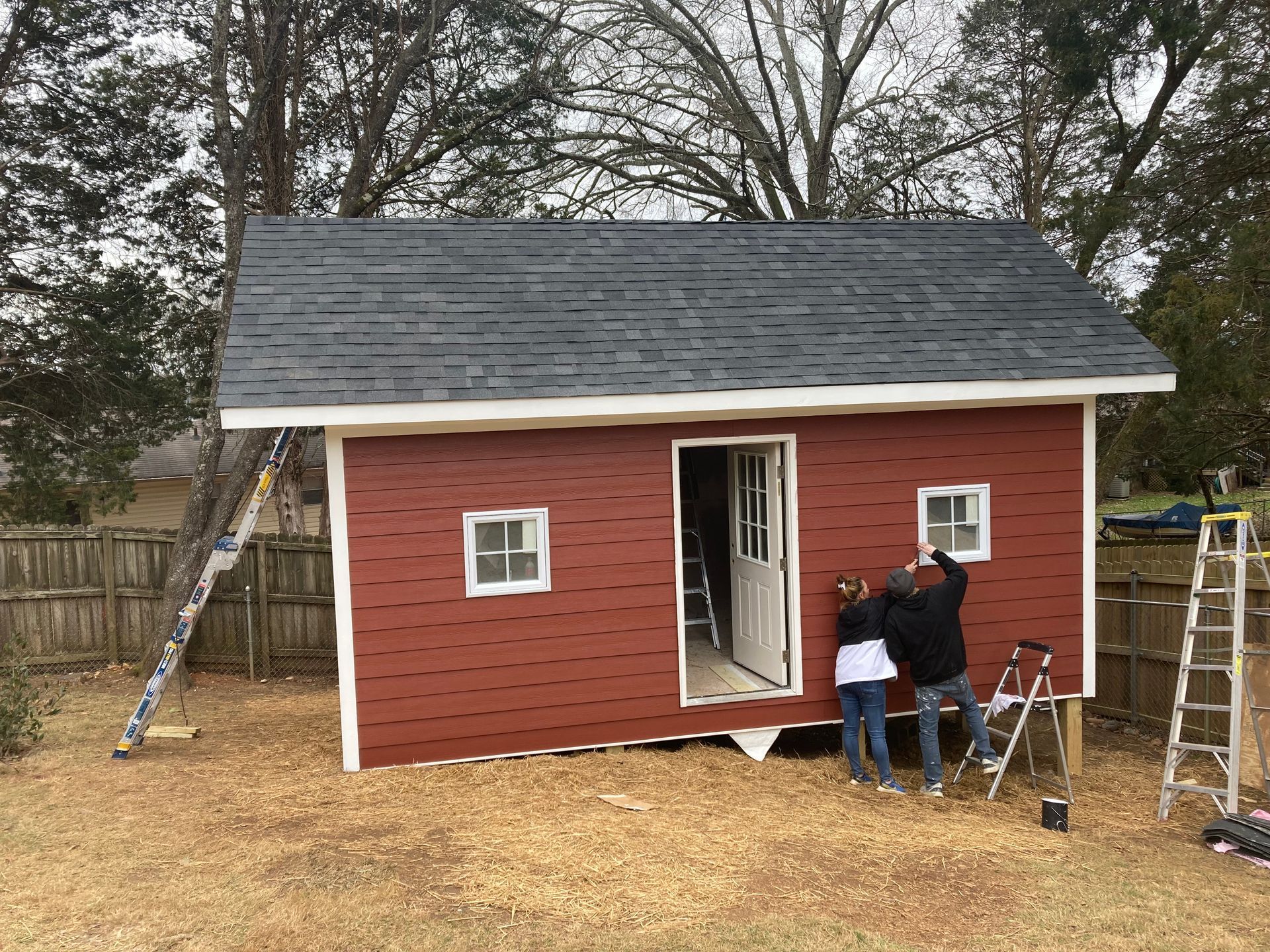 Red shed with a dark roof being worked on by two people. Wooden fence in background.