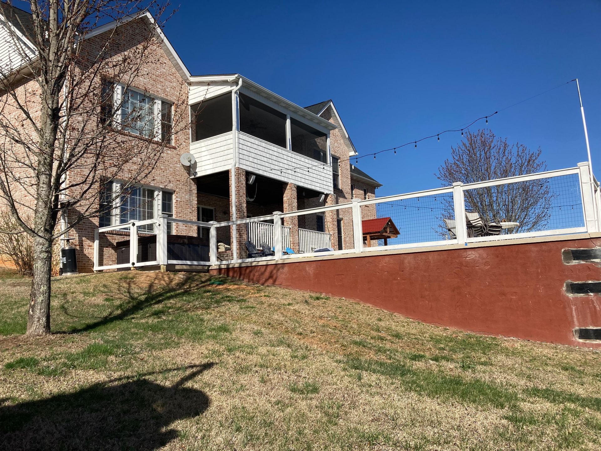Back of brick house with white railing and screened porch, over a red retaining wall on a grassy hill.