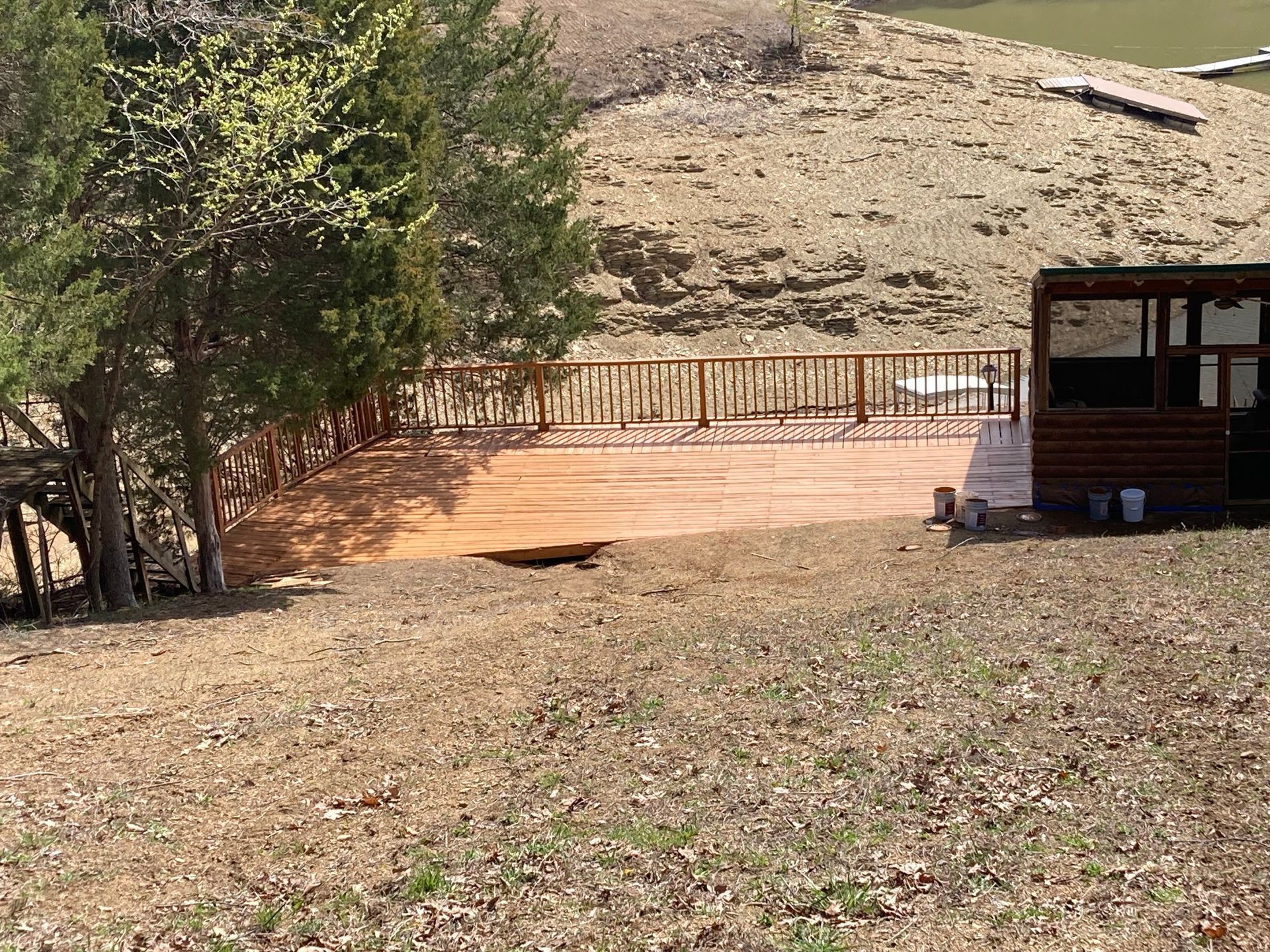 Wooden deck on a hillside with a small structure to the right, trees to the left, and a dry, grassy yard.