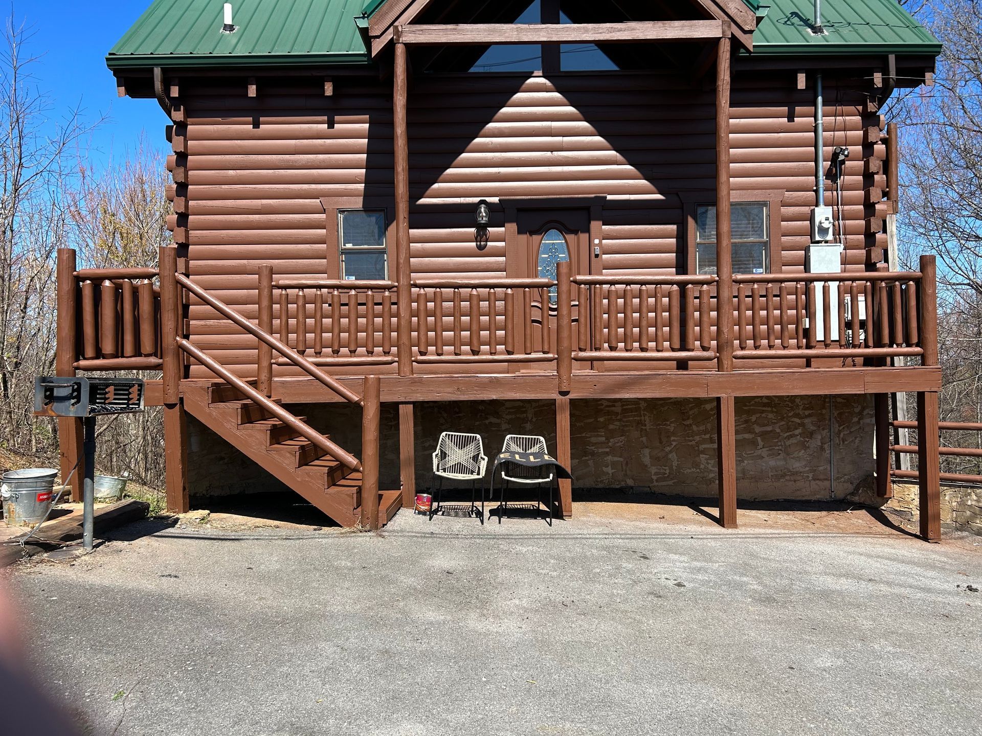 Brown log cabin with green roof and wooden deck, two chairs beneath.