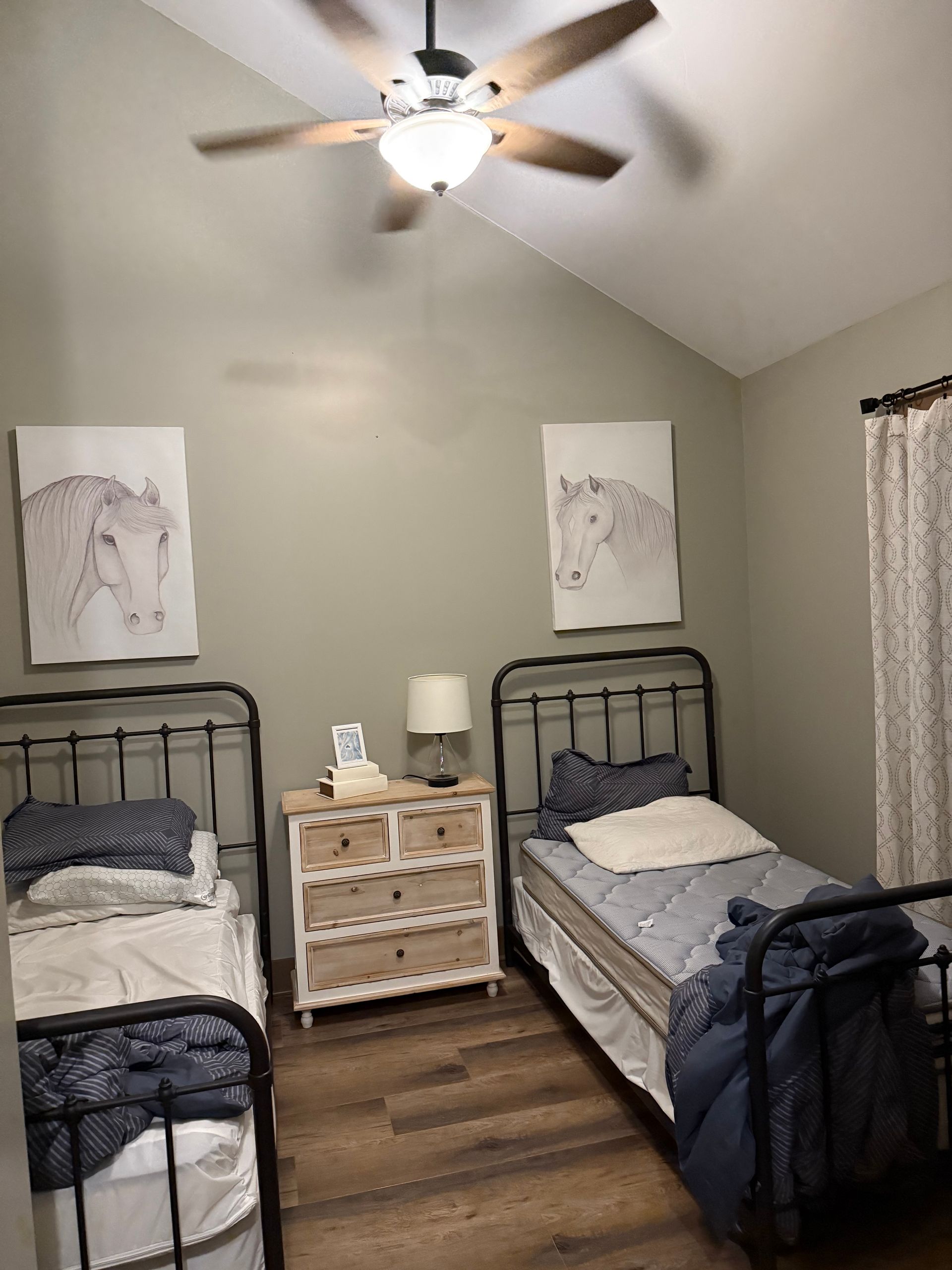 Two twin beds in a room with horse art, nightstand, and ceiling fan. Light-colored walls and wood-look flooring.