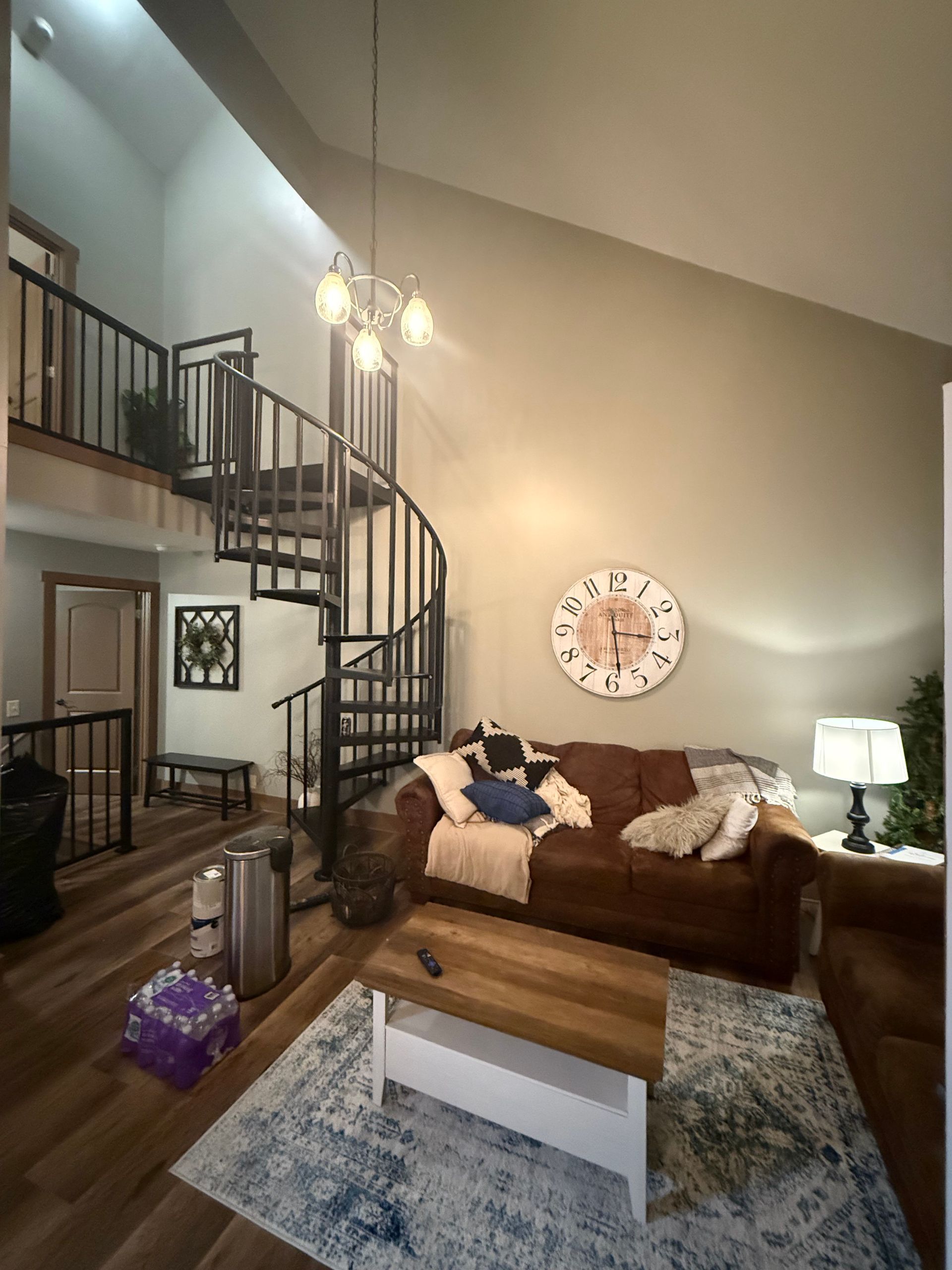 Living room with spiral staircase and brown sofa. Large clock, wooden coffee table, and blue rug.