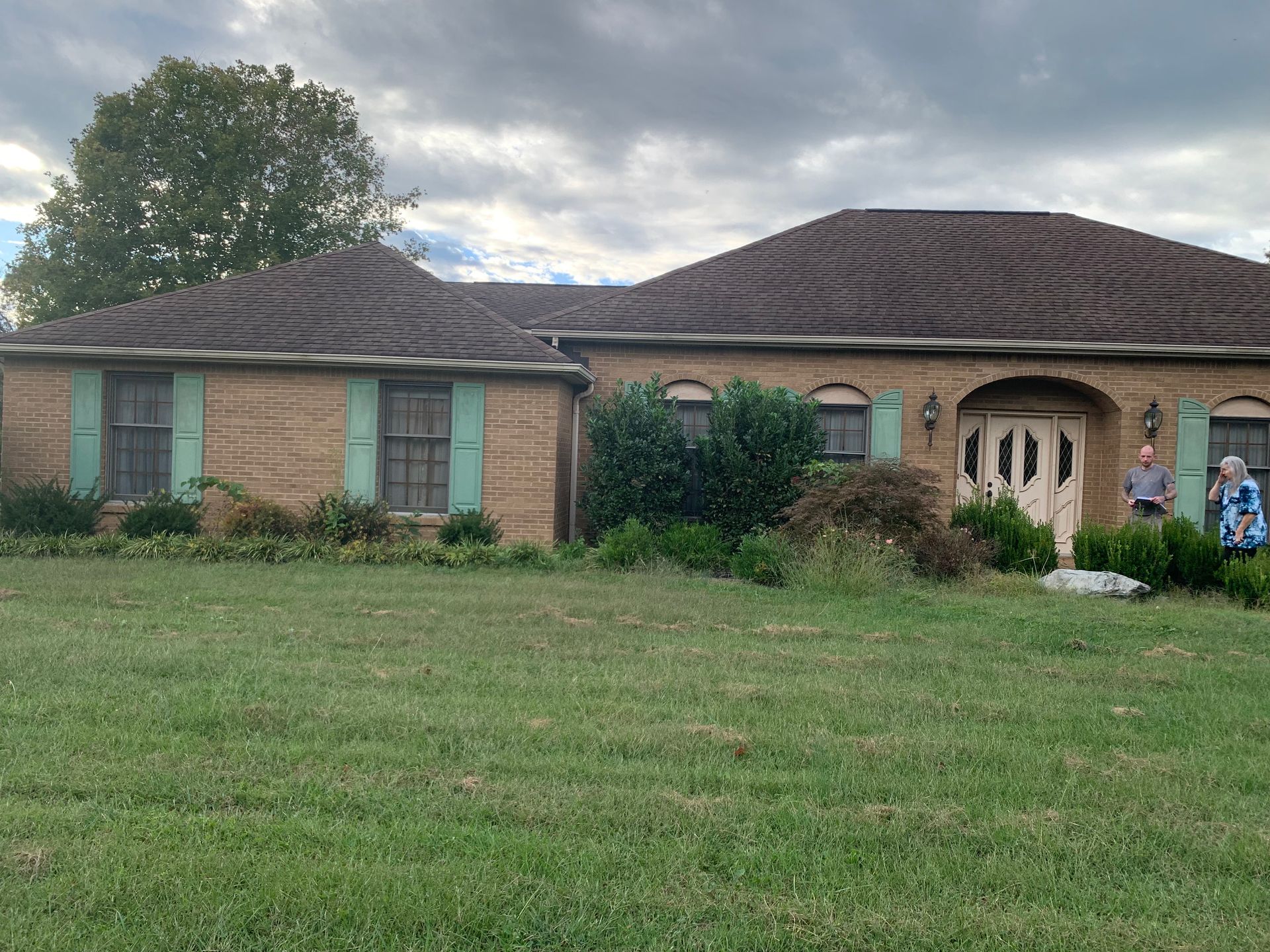 A tan brick house with brown roof and green shutters. Two people stand near the front door. Cloudy sky.