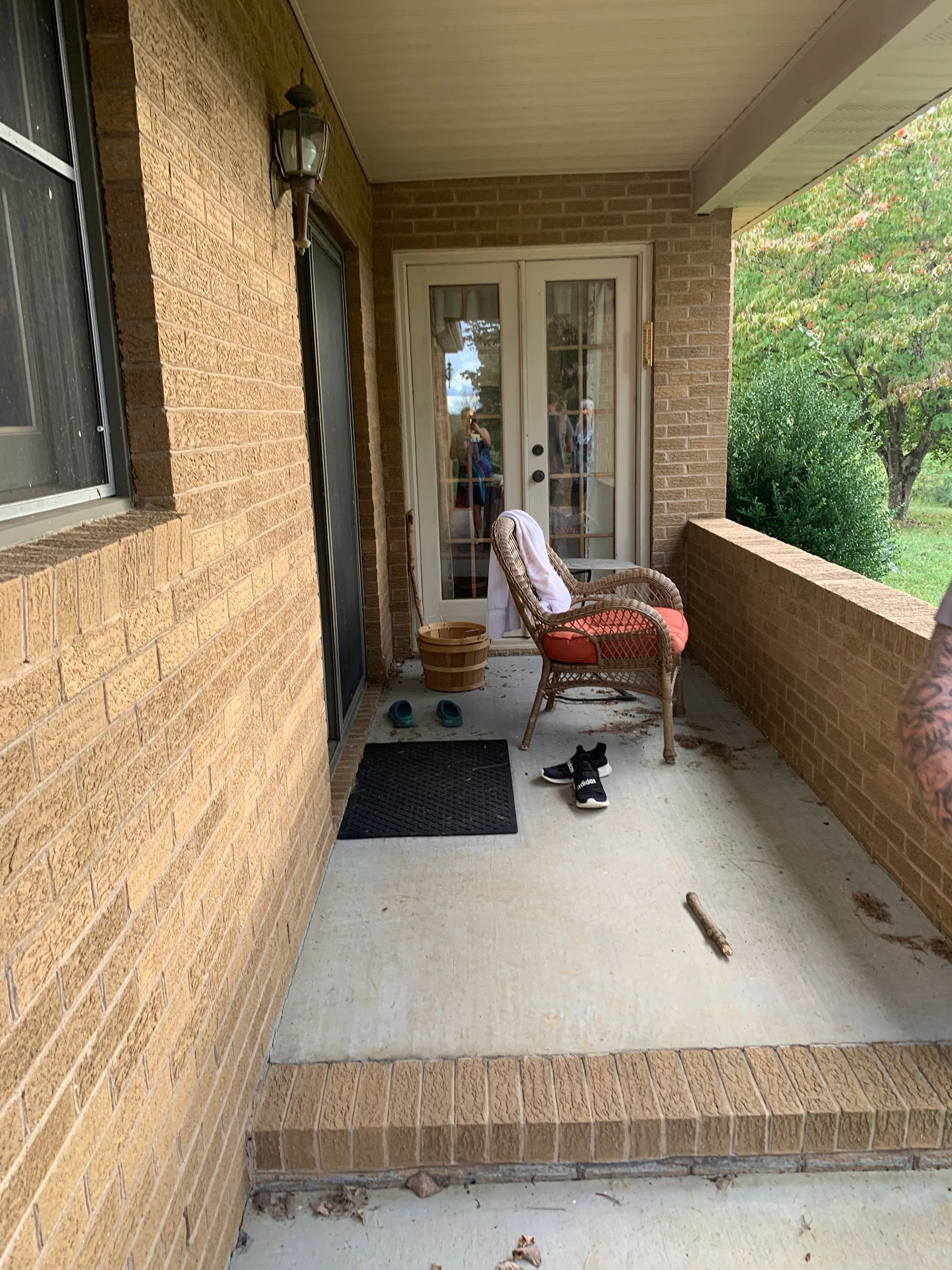 Covered porch with brick facade; wicker chair, doormat, and double doors lead inside.