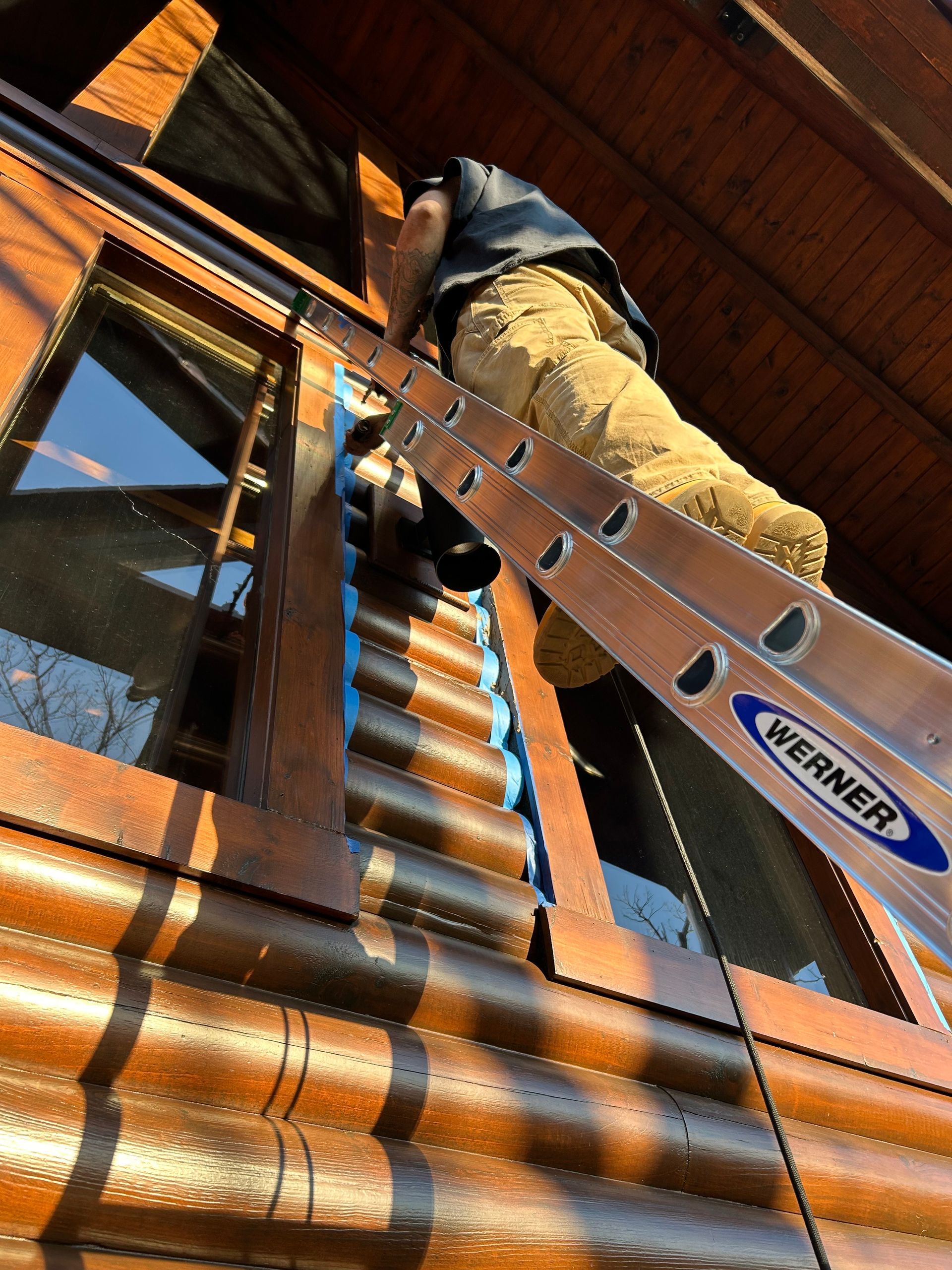 Man on ladder applying sealant to log cabin window frame.