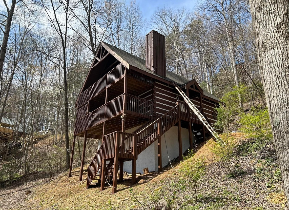 Brown log cabin with stairs and balcony on a hillside, surrounded by trees.