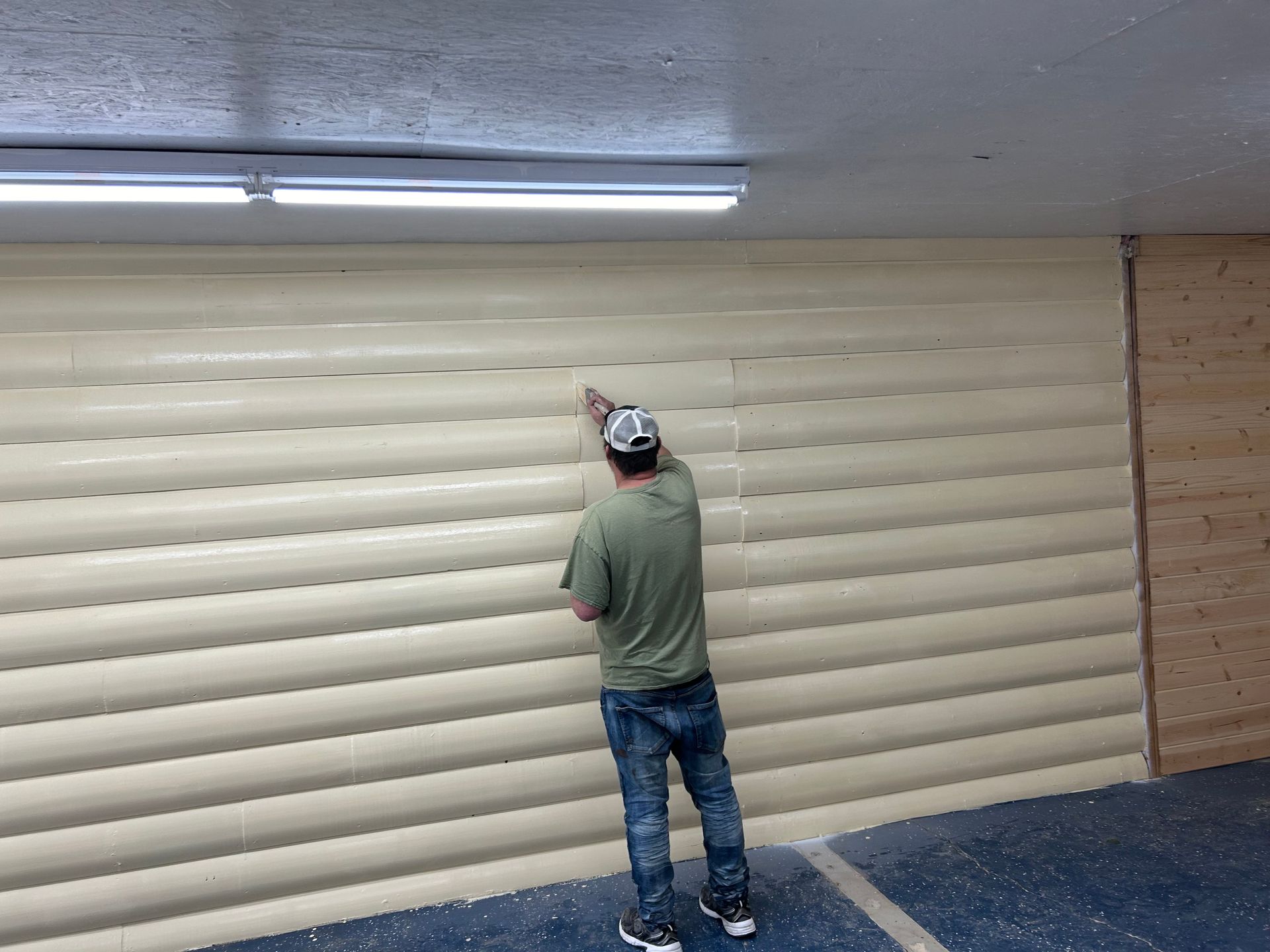 Man working on a tan, horizontal-paneled wall indoors, under a fluorescent light.