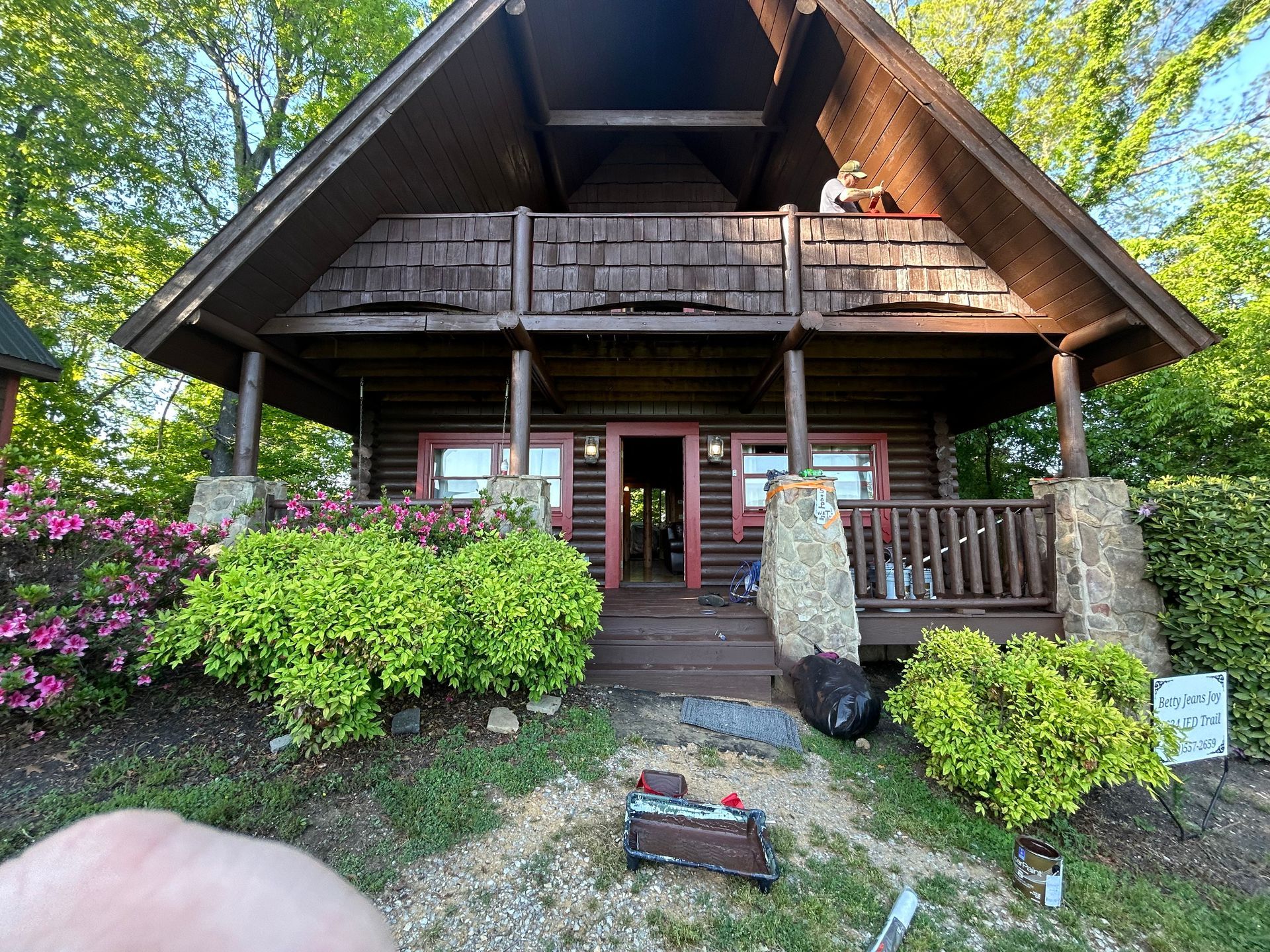 A-frame cabin with a porch. Brown exterior, green shrubs, and a person on the balcony.