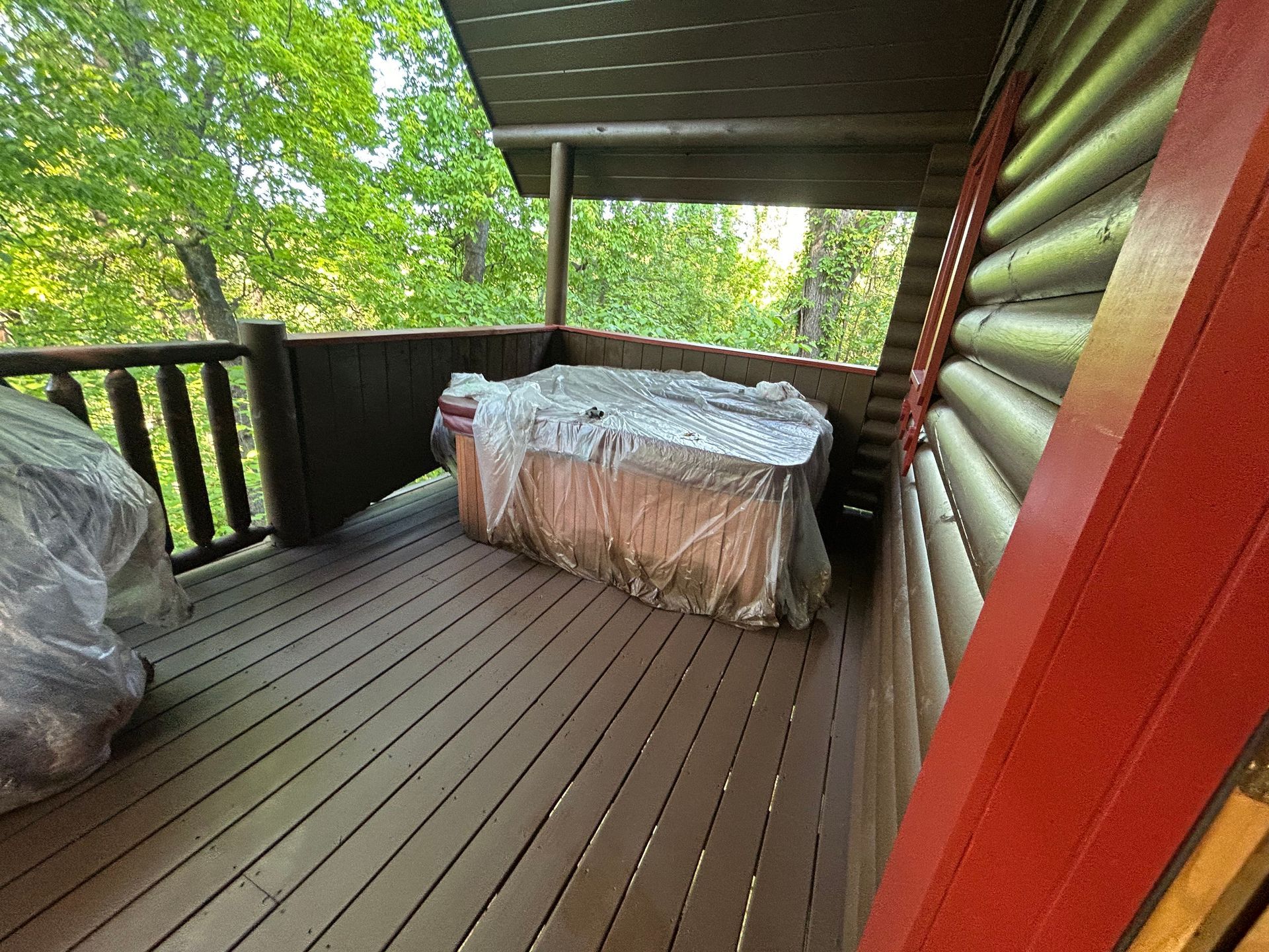 Covered hot tub on a wooden porch of a log cabin, trees in background.