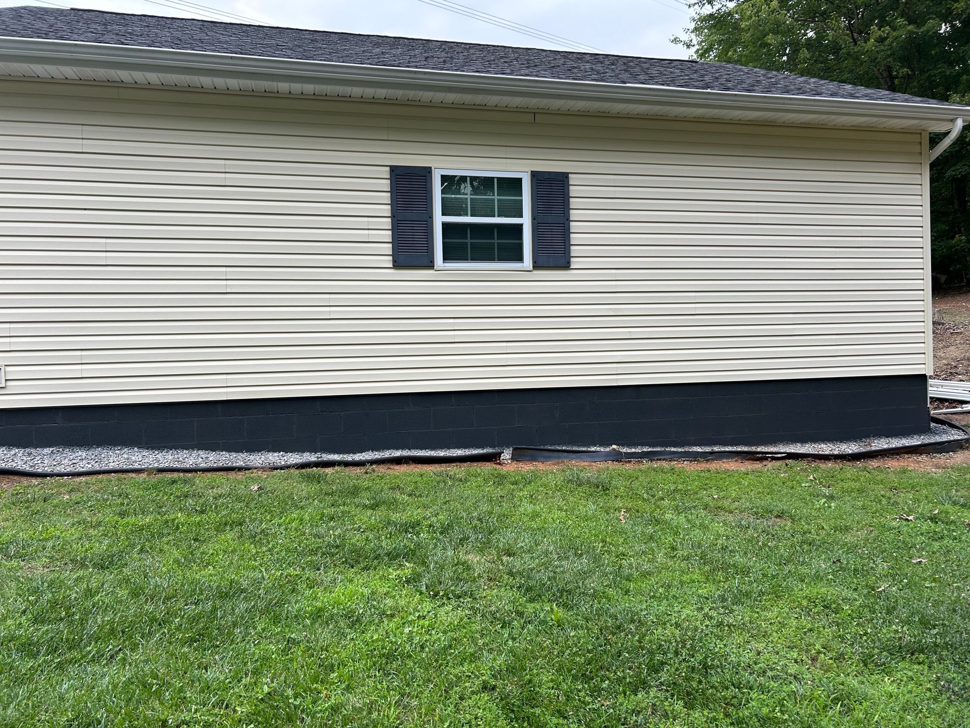 Beige siding on a building with a dark gray window shutter and a dark gray lower section, with green grass in front.