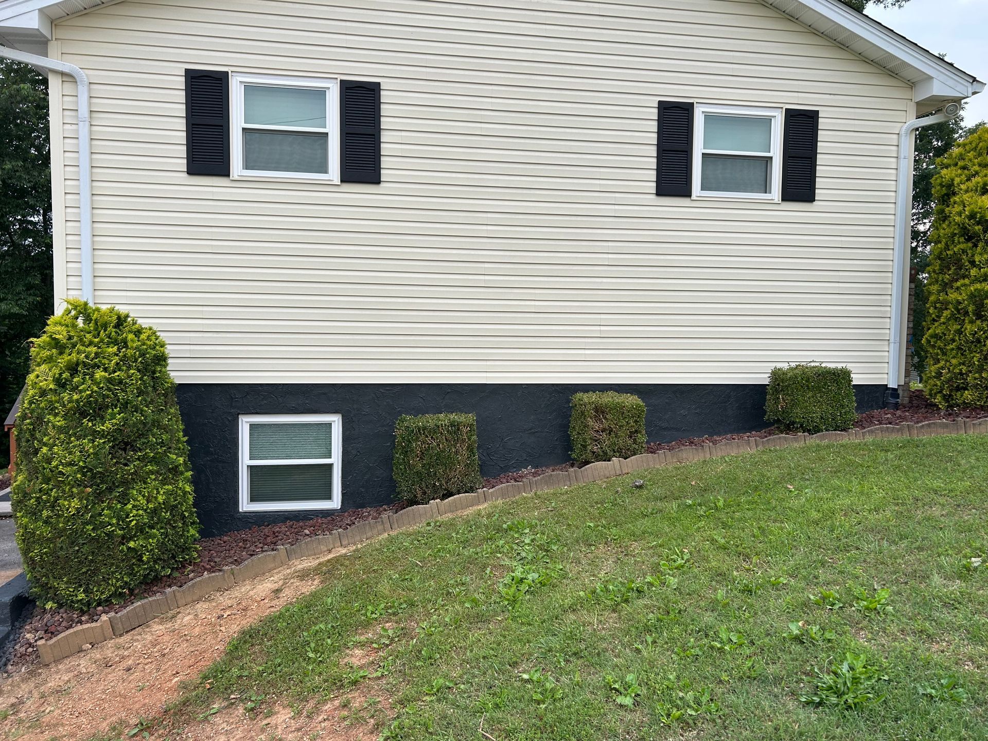 Beige house with black shutters, trim, and foundation. Green shrubs and grass on a slight slope.