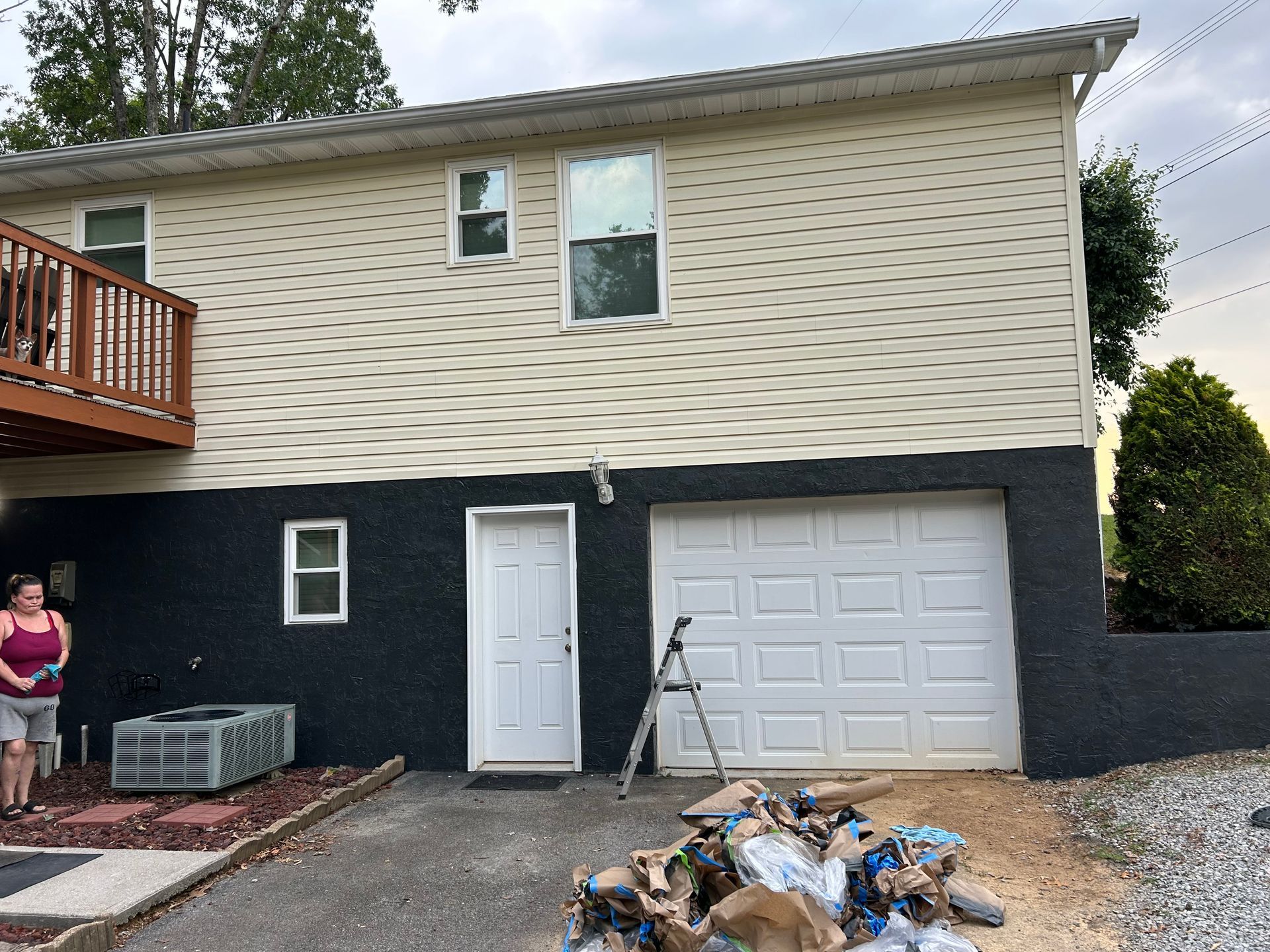 Side of a two-story house with beige siding above a dark blue/black base. White door and garage door. A person stands nearby.