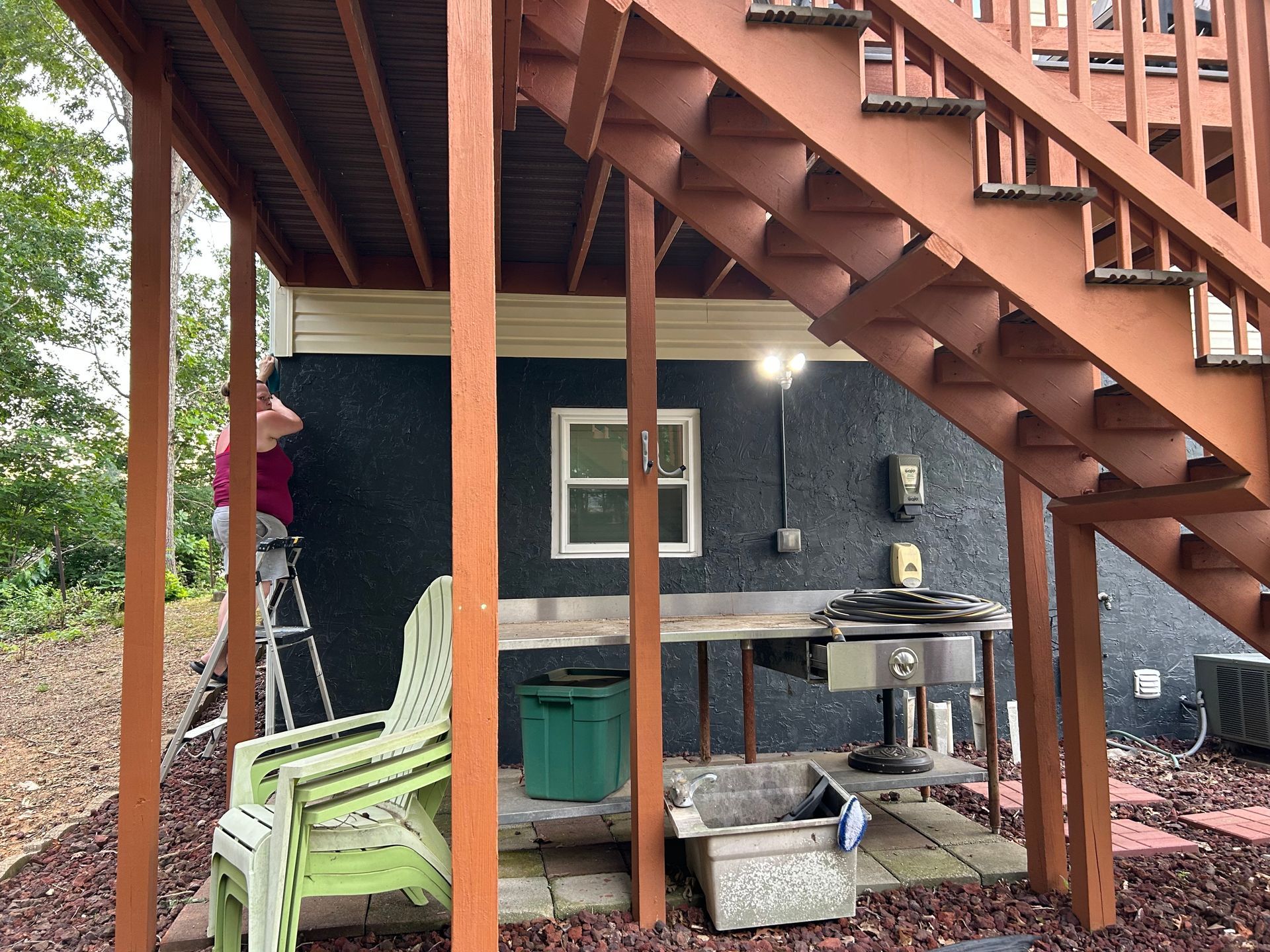 Brown deck stairs, supporting beams, exterior grill setup under structure; person on ladder.