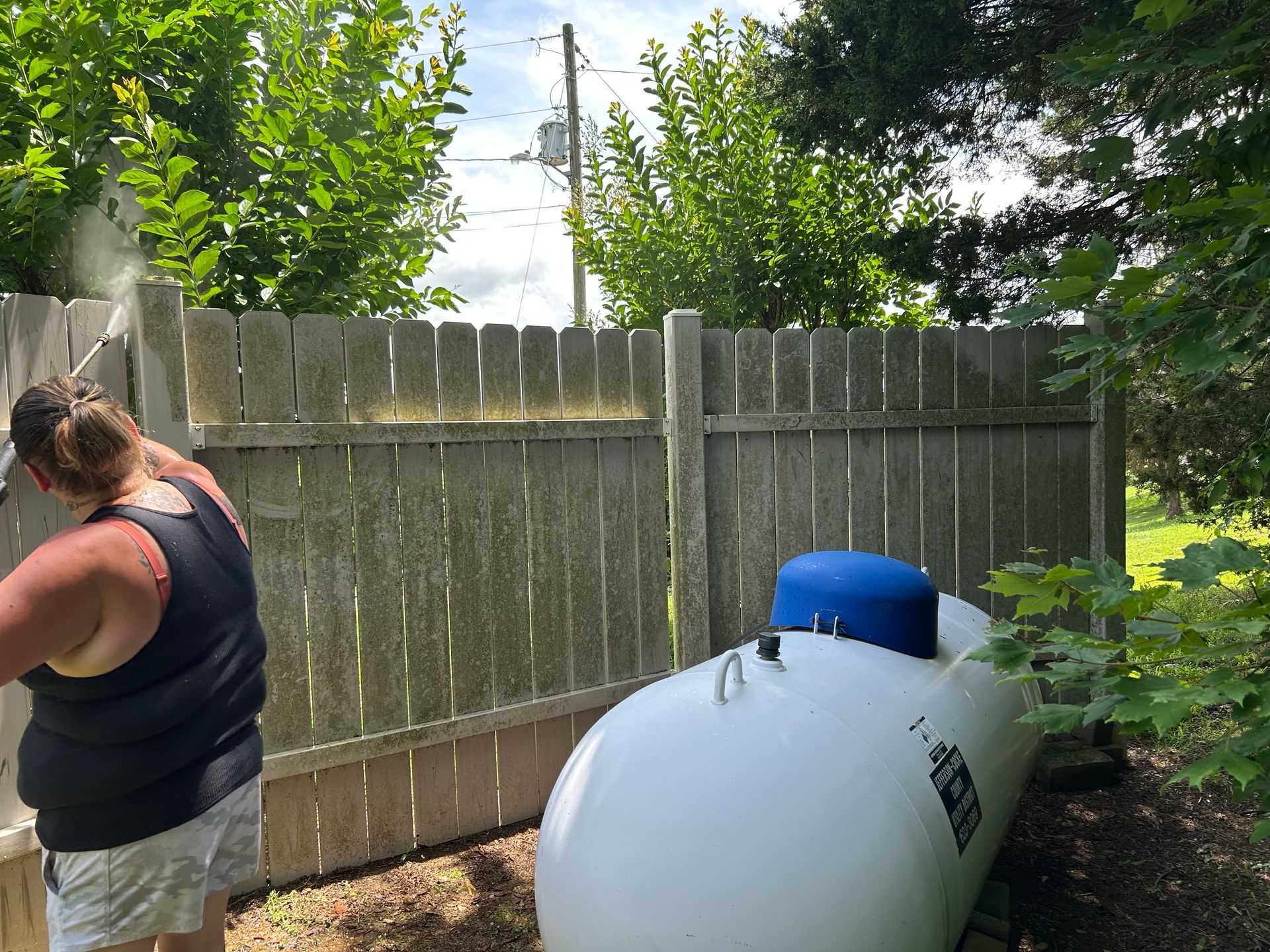 Person pressure washing a wooden fence next to a large white propane tank. Green trees and a cloudy sky in background.