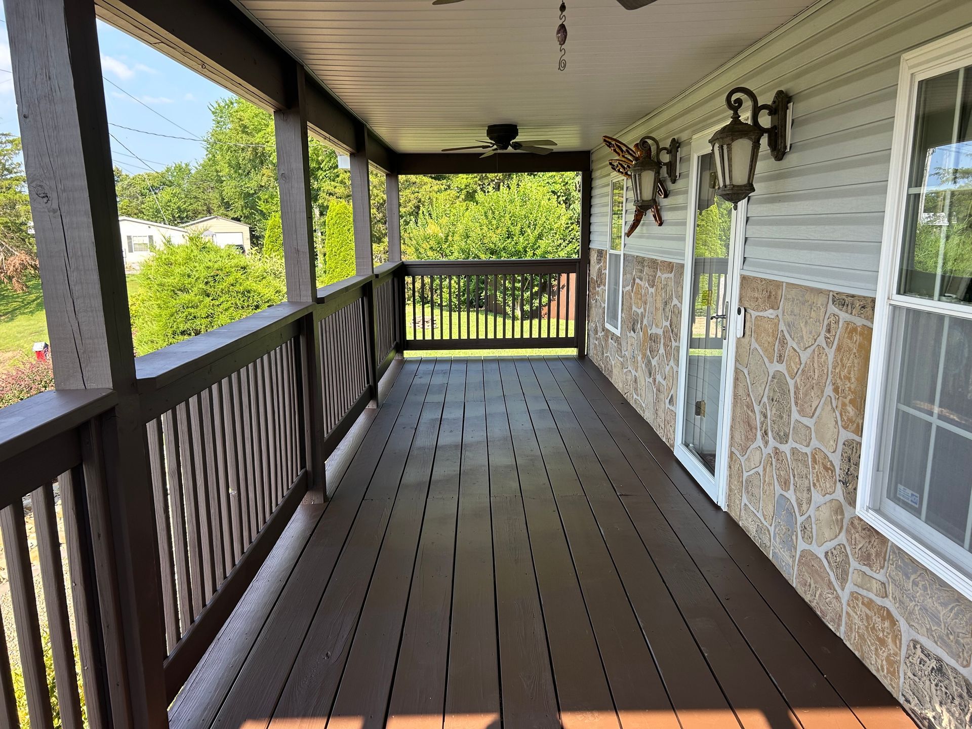 Covered porch with brown flooring, railings, and ceiling fan. Stone and vinyl siding on the house.