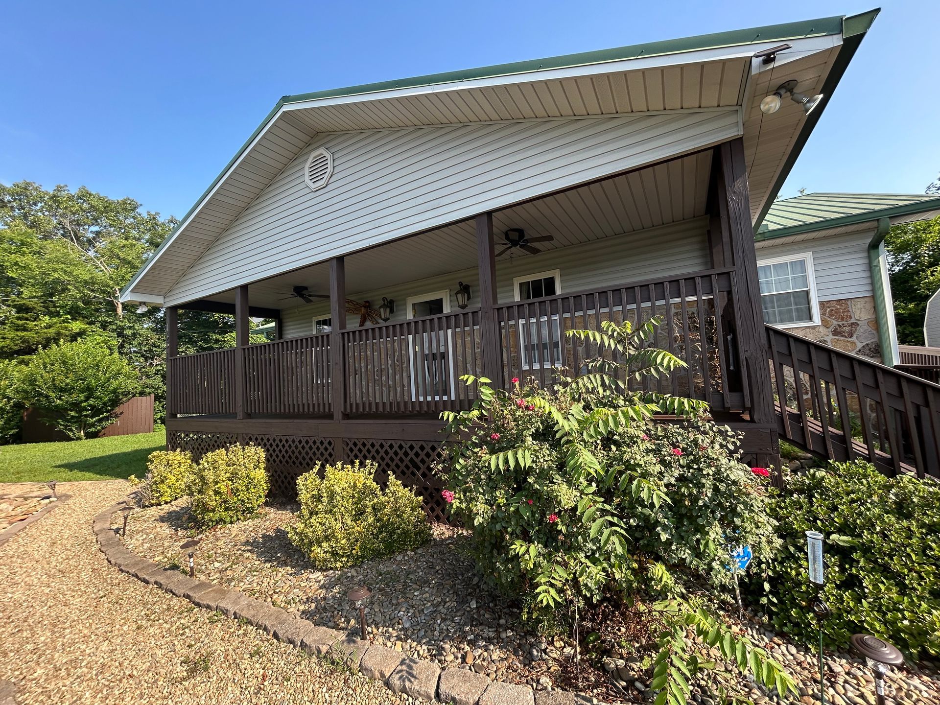 Cabin with brown porch and green roof, surrounded by bushes and trees on a sunny day.