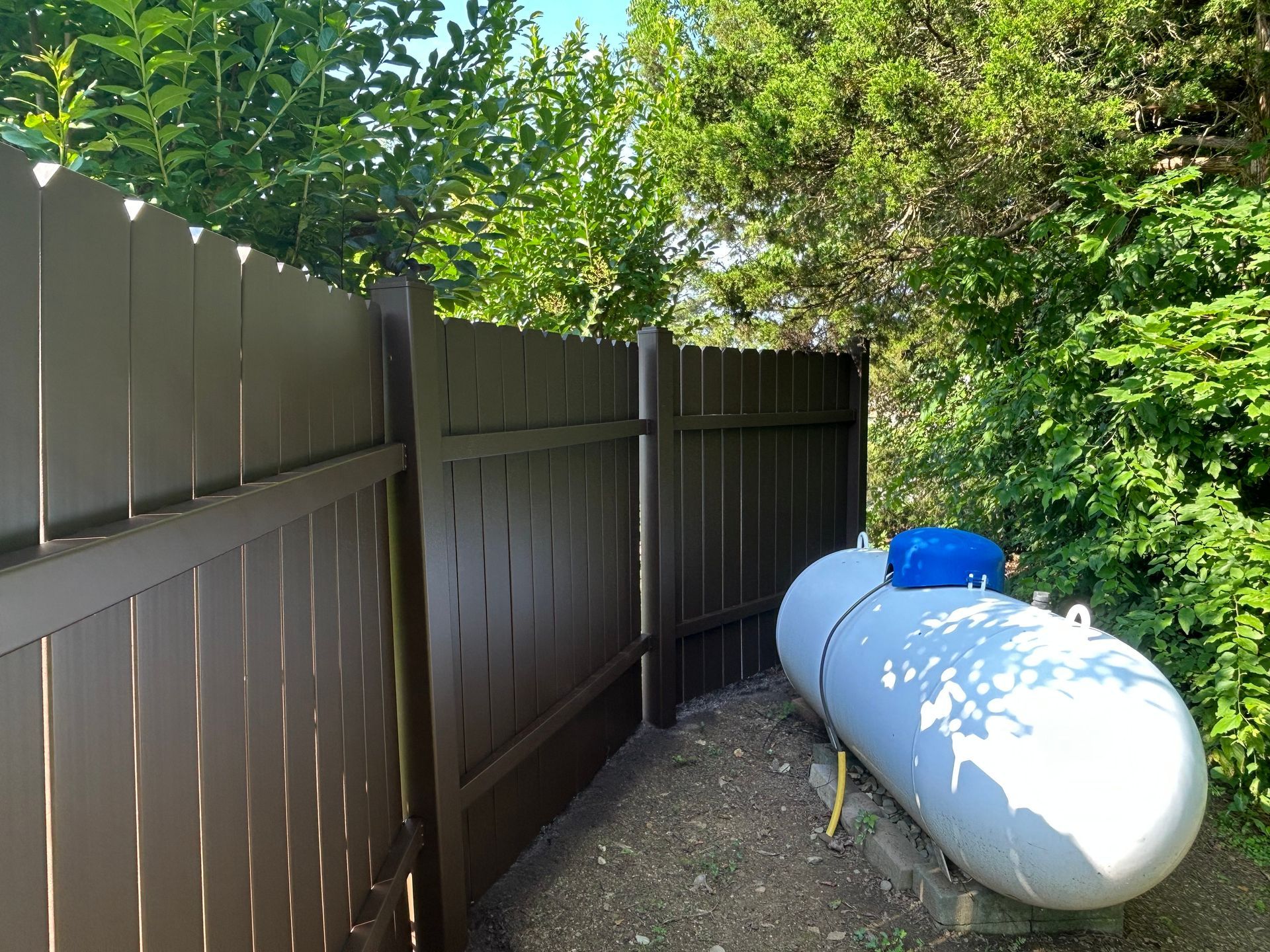 Brown fence surrounding a large propane tank in a yard with green trees.