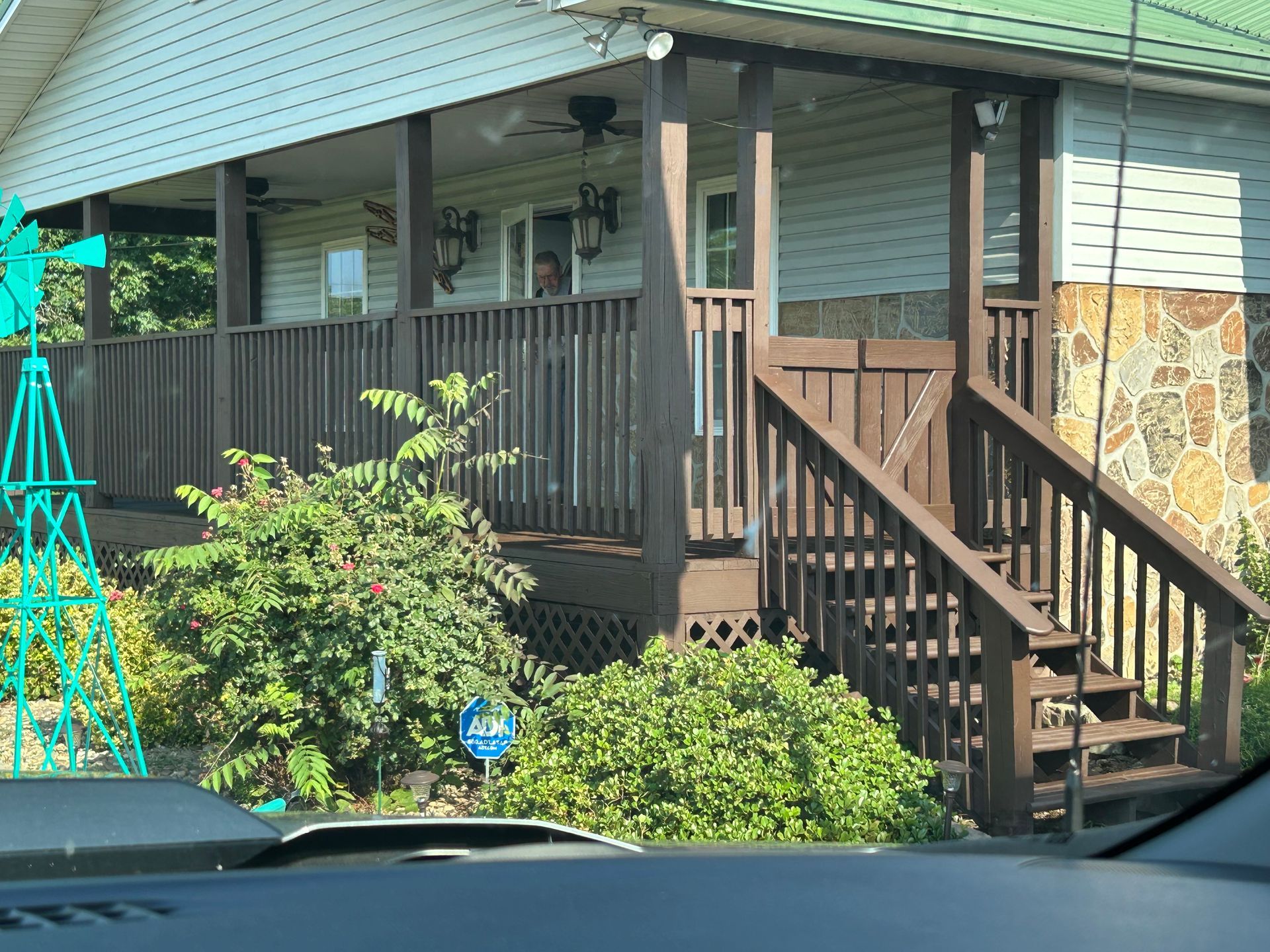 House with a wraparound porch, wooden railing and stairs. Green and brown exterior, stone accent.