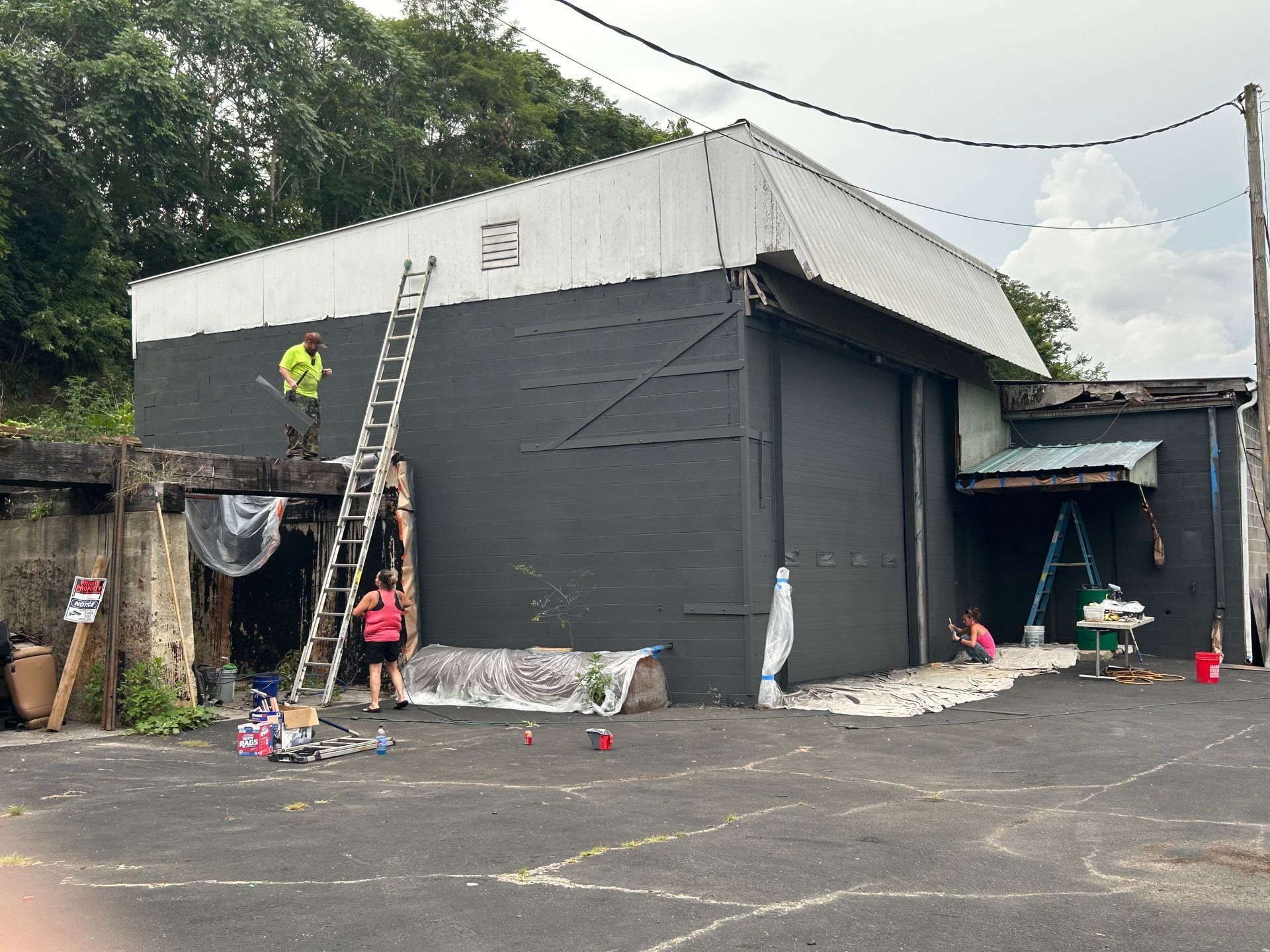 People painting a dark building; one on a ladder, two on the ground. Exterior scene, sunny day.
