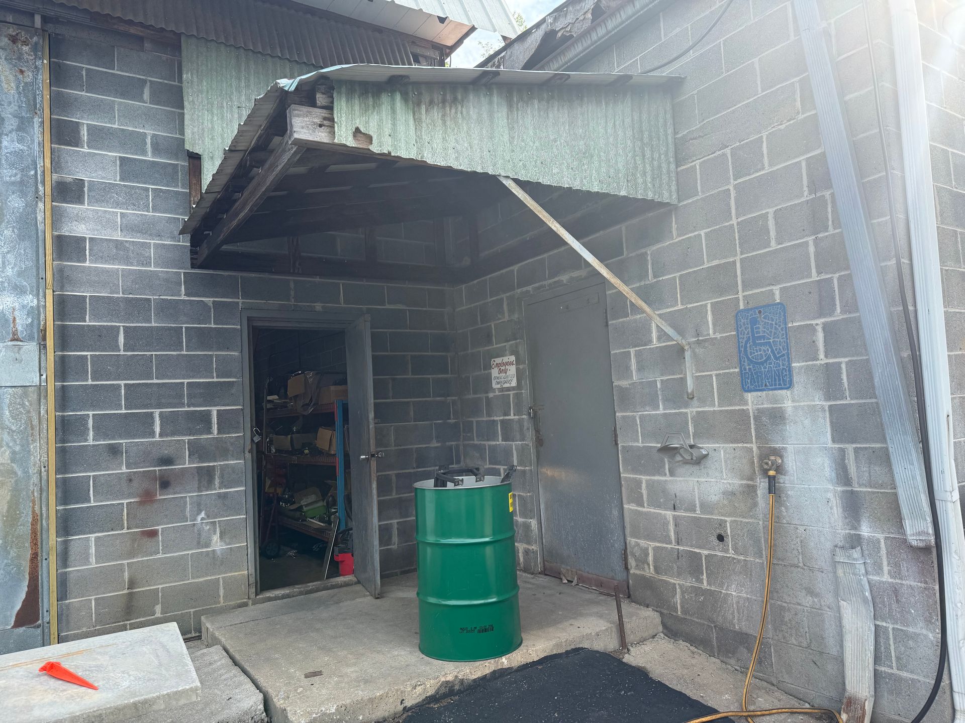 Exterior of a gray brick building with a metal awning. A green barrel sits below the awning next to a door.