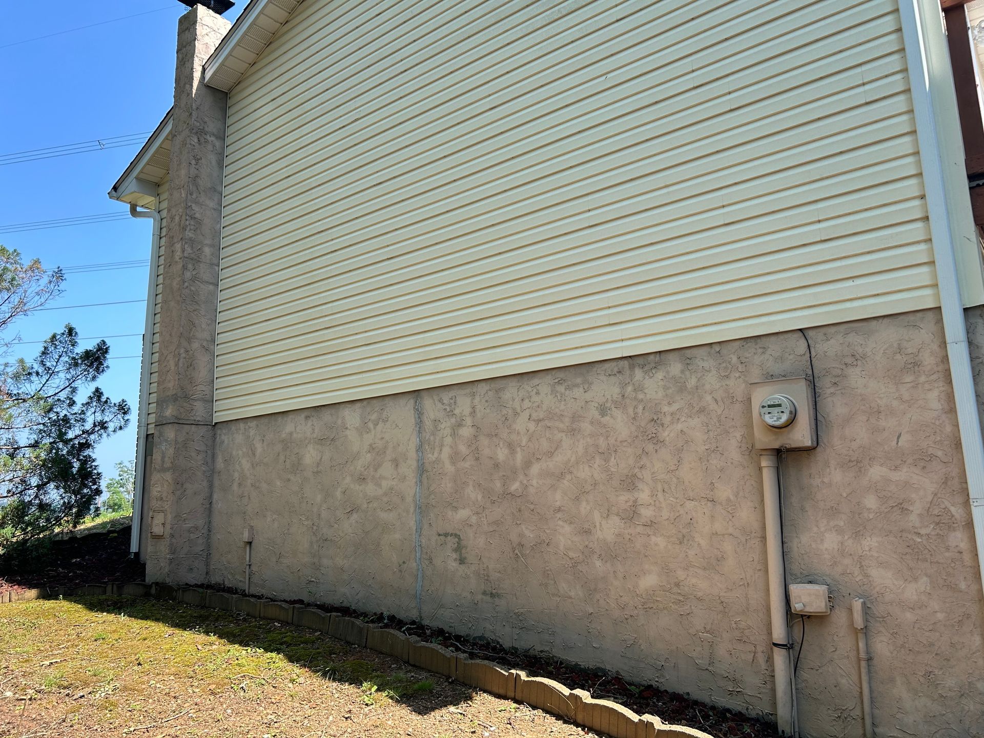 Side view of a house with beige siding and stucco base, electrical box visible. Sunny day.