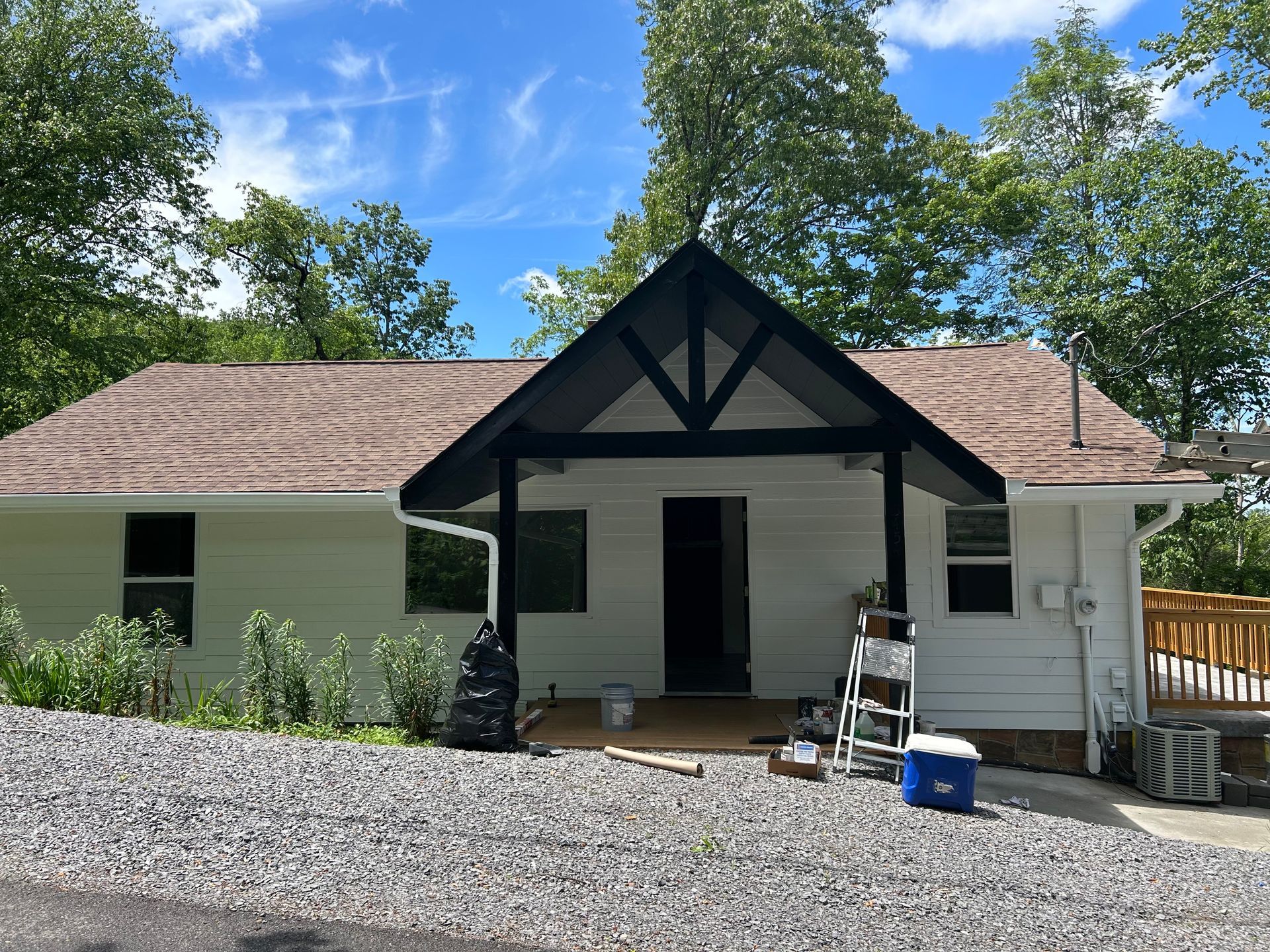 A white house with brown roof and black porch under a blue sky. Gravel driveway and bushes in front.